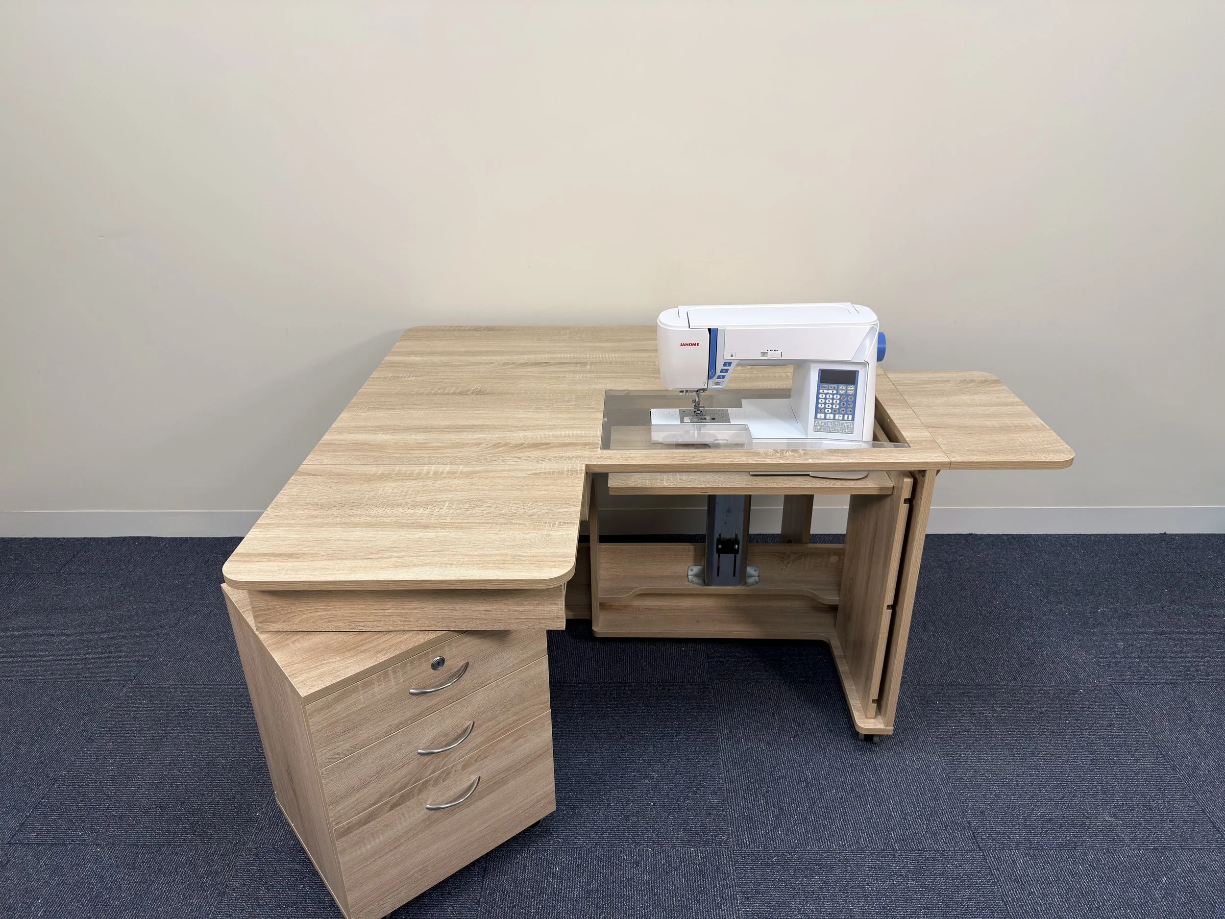 Wooden sewing or embroidery machine table with a white Janome machine on top, and a set of three drawers on the left side, placed on a dark carpeted floor against a plain light-colored wall.