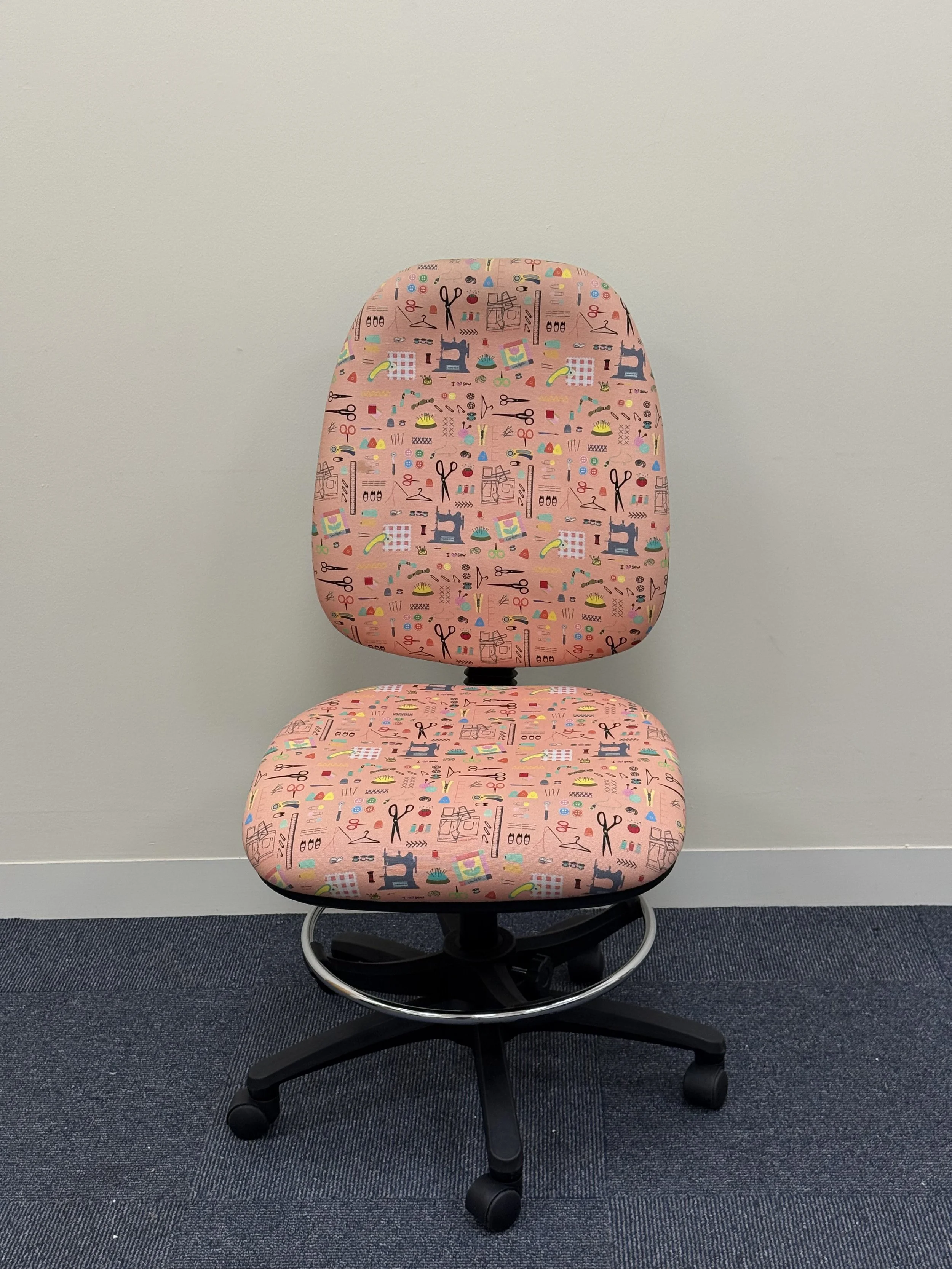 Office chair with pink fabric featuring sewing-themed illustrations, positioned against a plain light-colored wall.