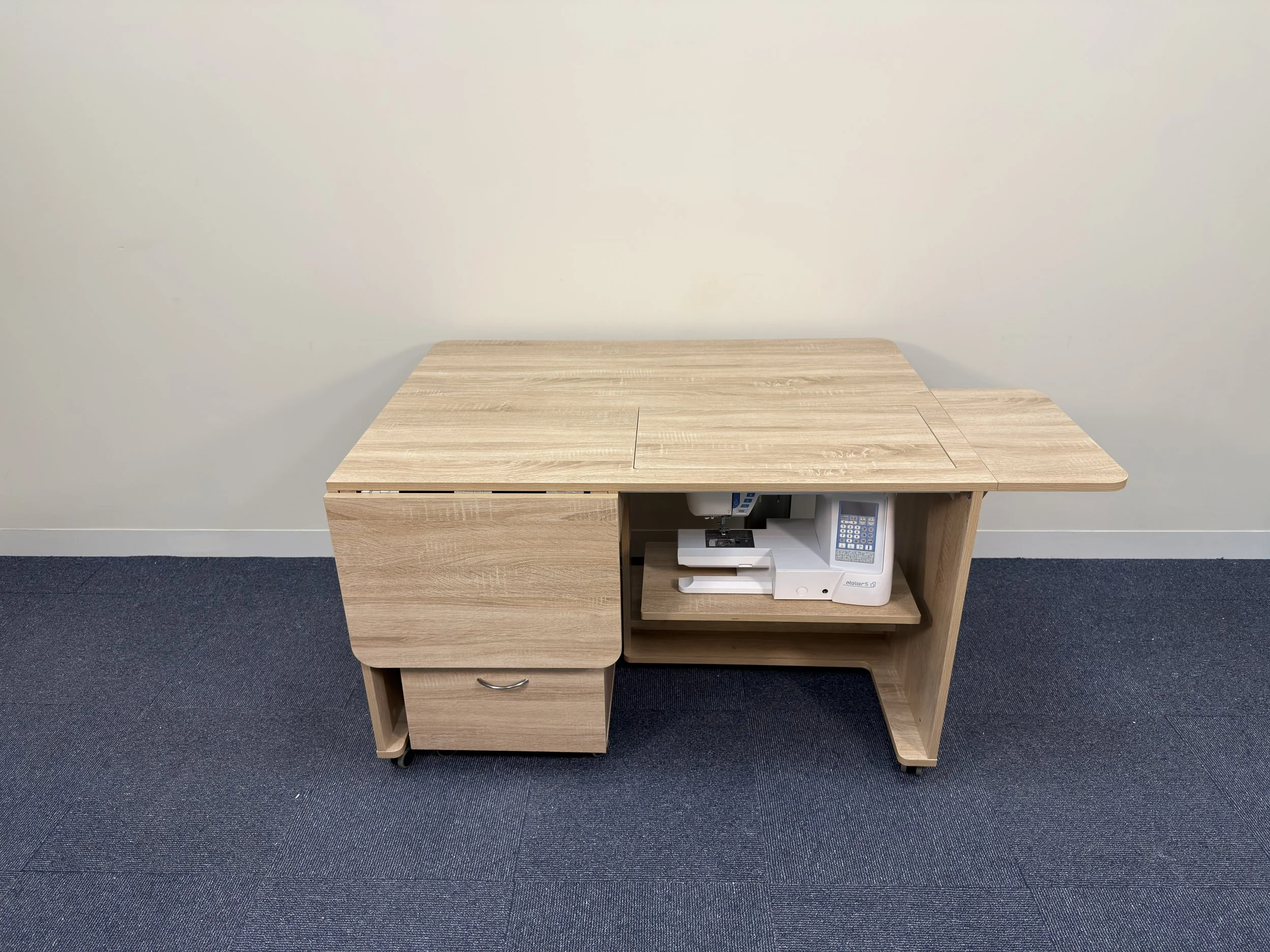 Light wood office desk with a sliding top, a drawer on the left, and a built-in shelf on the right holding a telephone and a notepad. The desk is against a plain white wall with a dark blue carpeted floor.