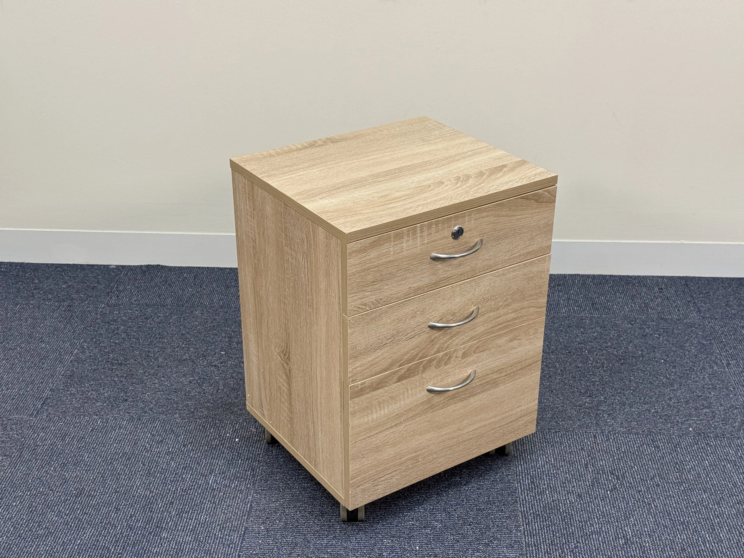 A light wood filing cabinet with three drawers, each with a silver handle, placed on a dark blue carpeted floor against a plain white wall.