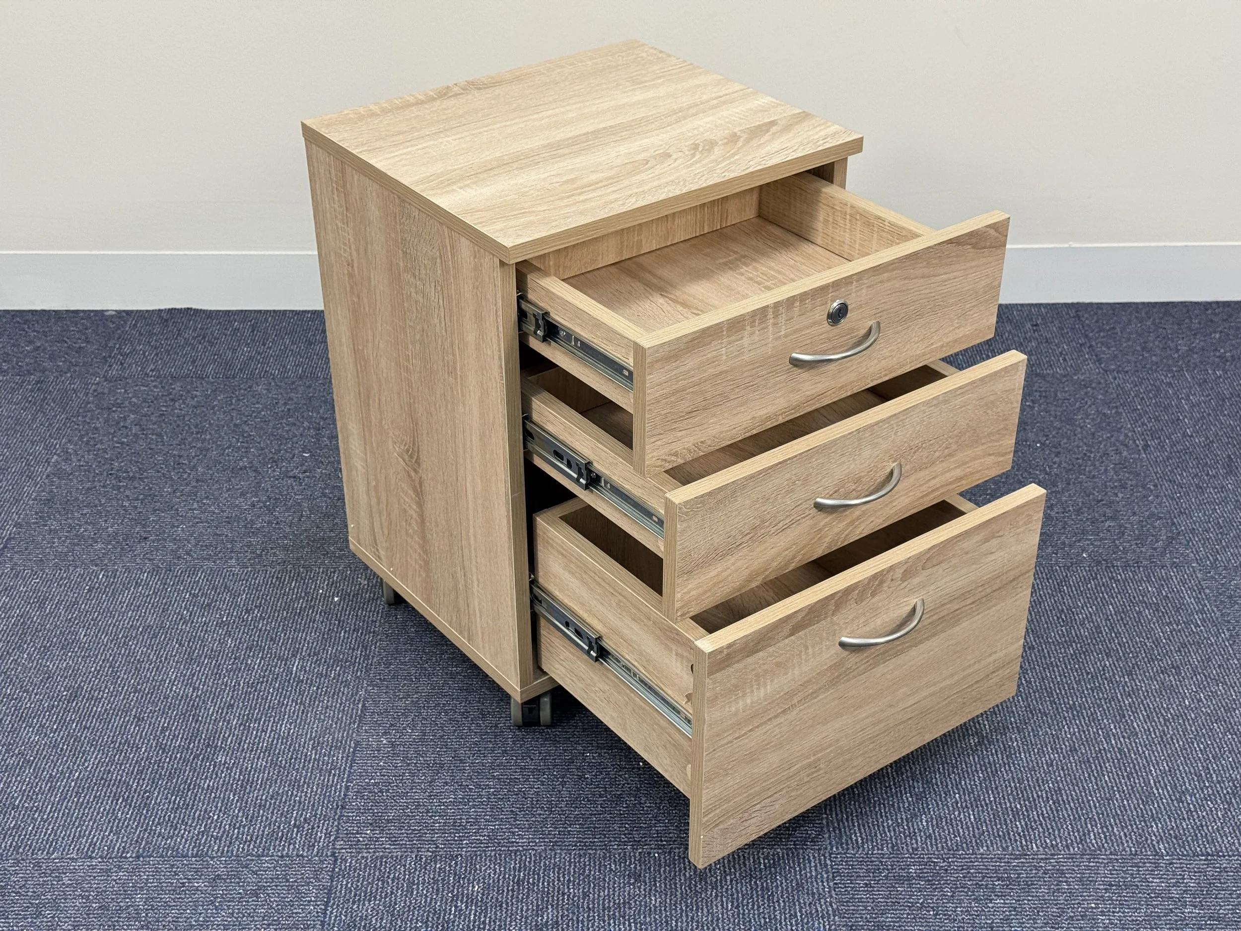 A light wood three-drawer filing cabinet with silver handles and metal rails, placed on a blue carpeted floor against a beige wall.