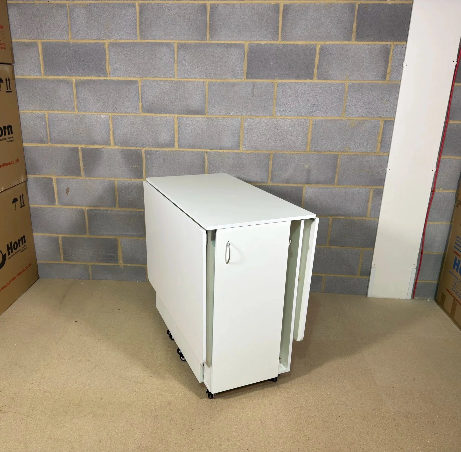 A white, foldable table or cart on wheels, partially folded, in a room with a concrete block wall and some boxes.