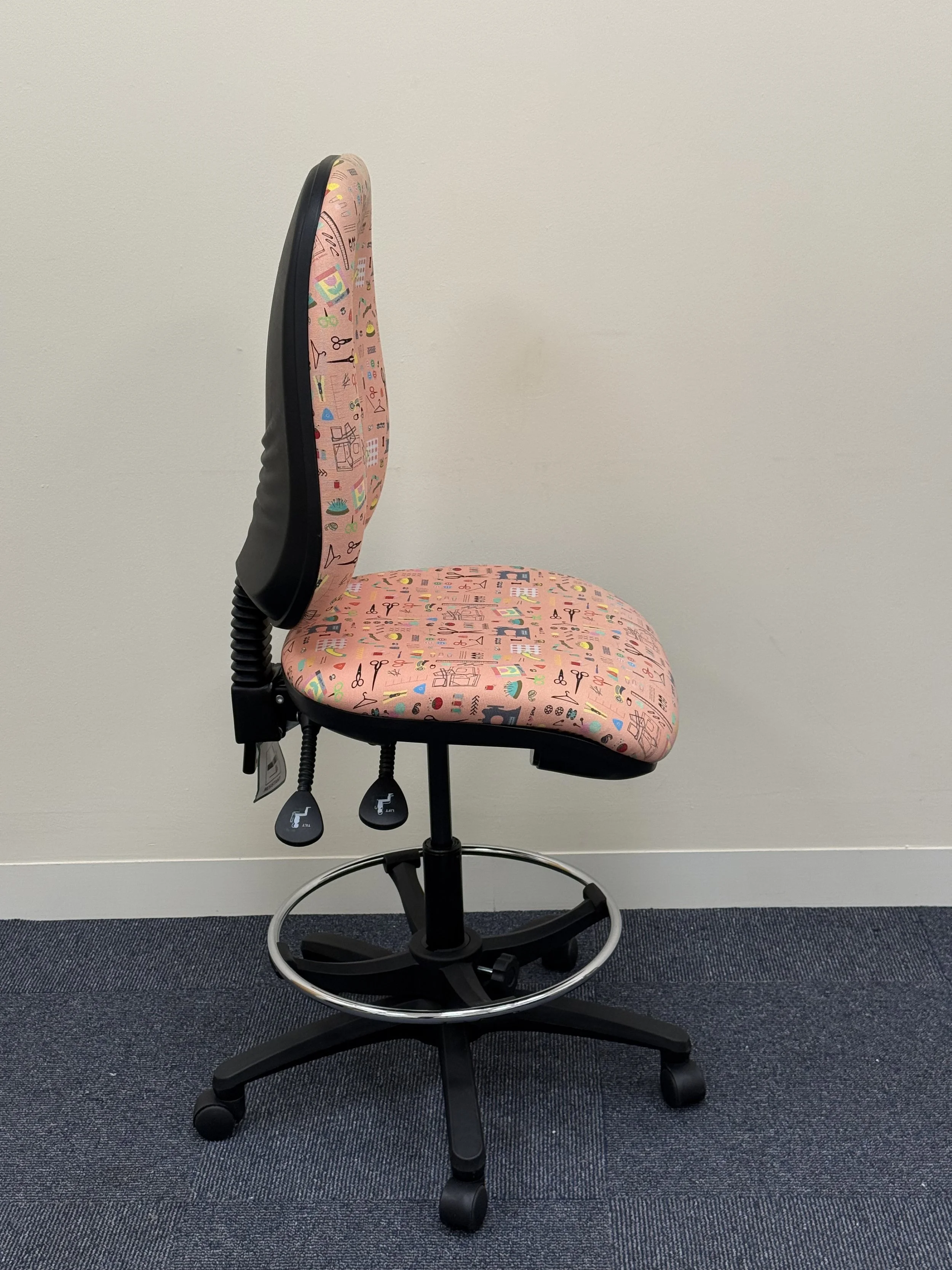 Office chair with pink fabric upholstery featuring colorful abstract doodles, black armrests, adjustable height levers, a metal footrest ring, and black wheeled base, positioned against a plain white wall on dark blue carpet.