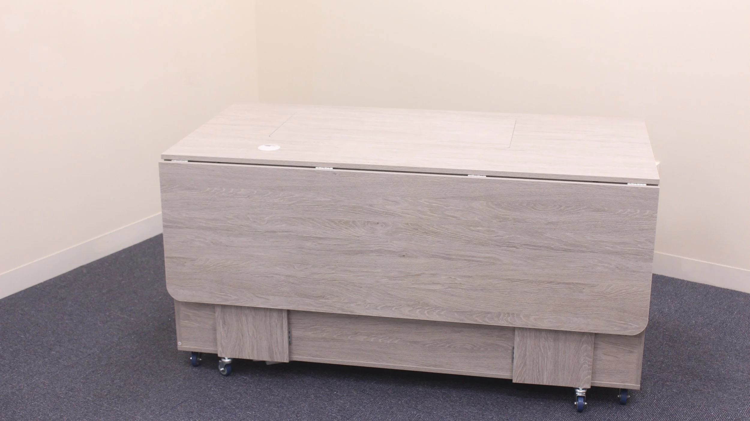 A light wood-colored rectangular storage chest on wheels in a room with plain beige walls and dark gray carpet.