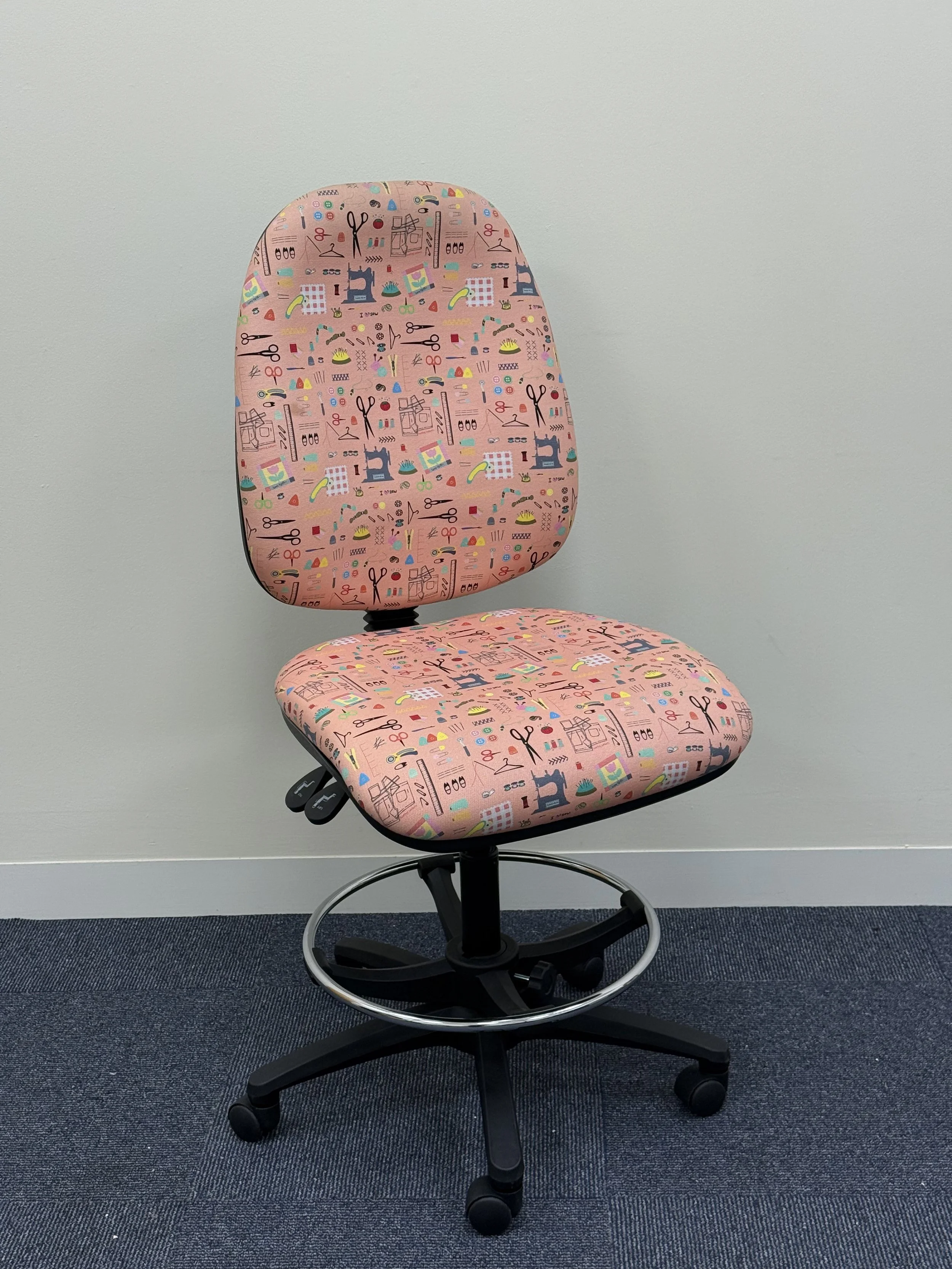 Office chair with pink fabric featuring sewing, scissors, thread, and fabric patterns, positioned against a plain white wall and blue carpeted floor.