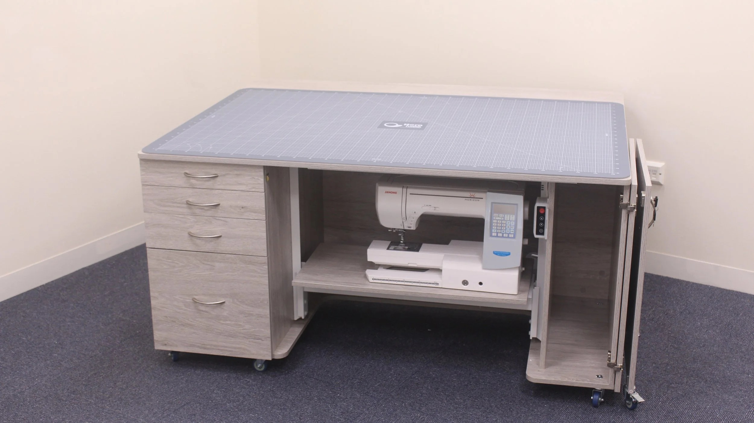 A sewing machine placed inside a wooden crafting table with a cutting mat on top, in a room with beige walls and dark carpeted floor.