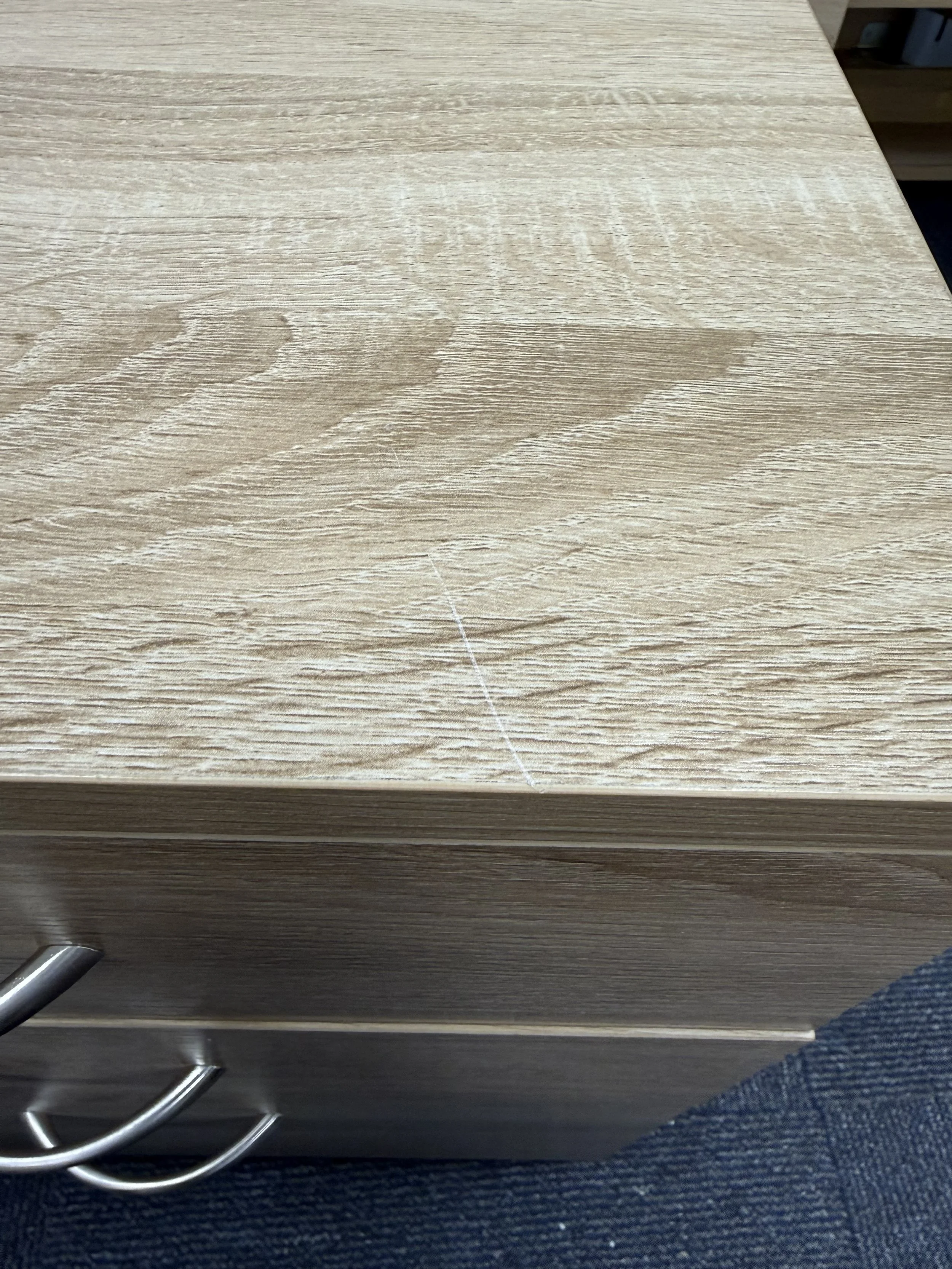 A light wood-colored drawer with a scratch on the top surface, showing a horizontal metal handle and a blue fabric in the lower right corner.