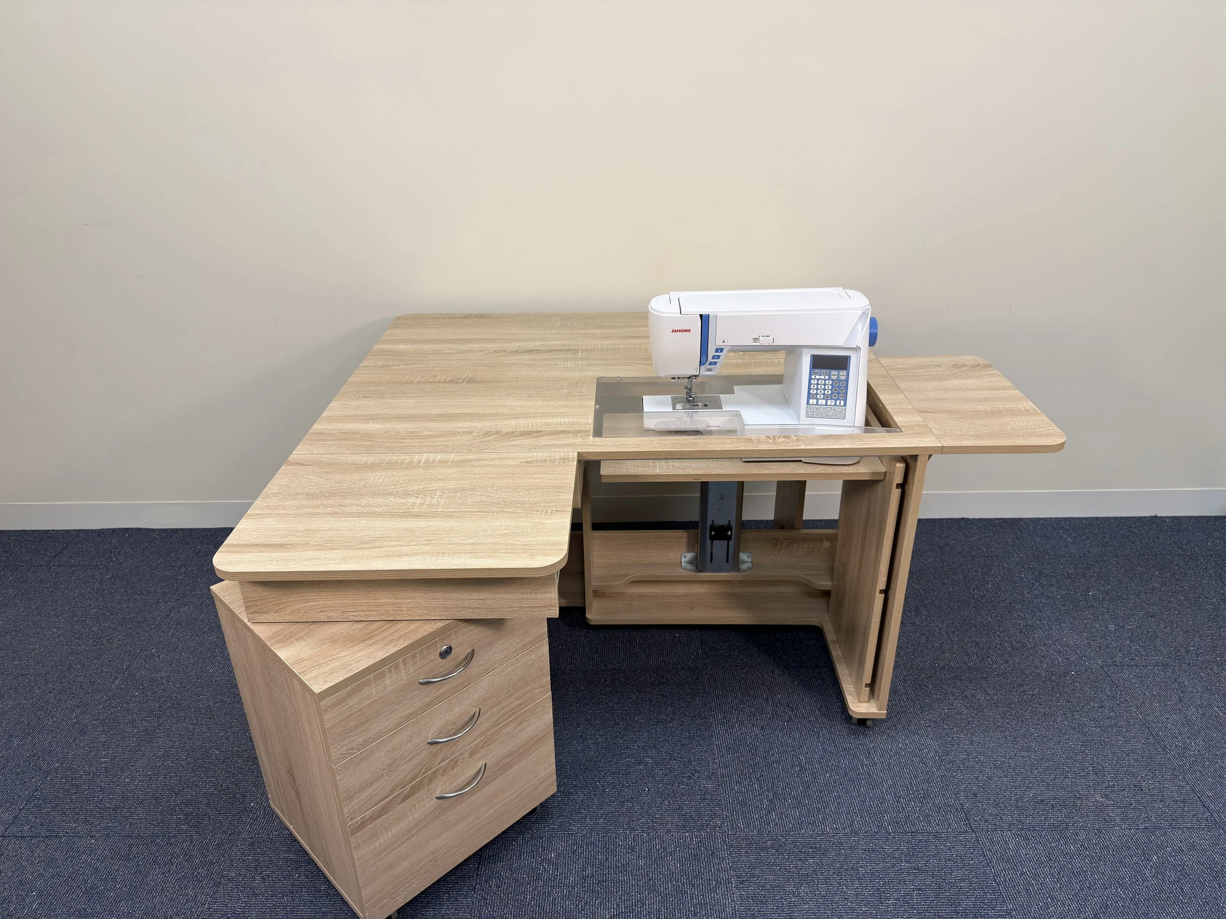 A wooden desk with a sewing machine on top, positioned in an office or craft room with beige walls and dark gray carpet.