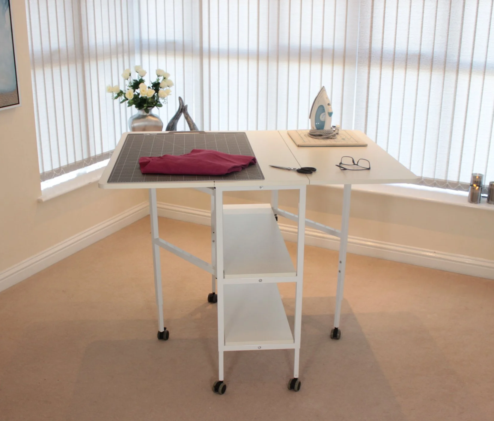 White ironing board with a pink cloth, an iron, scissors, and glasses on top, placed on beige carpet in a room with large bay windows.