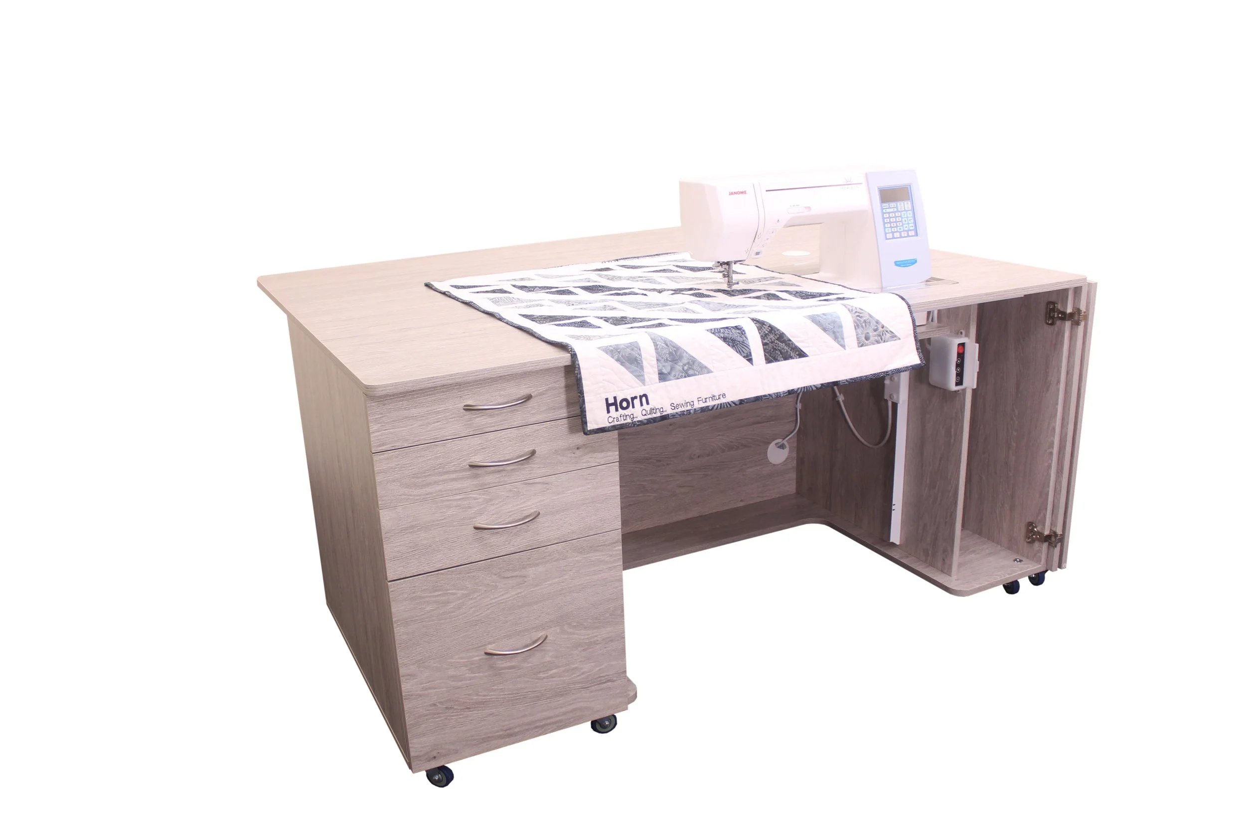 A sewing machine on a wooden desk with open storage cabinet, partially covered by a quilted fabric projects, with a white background.