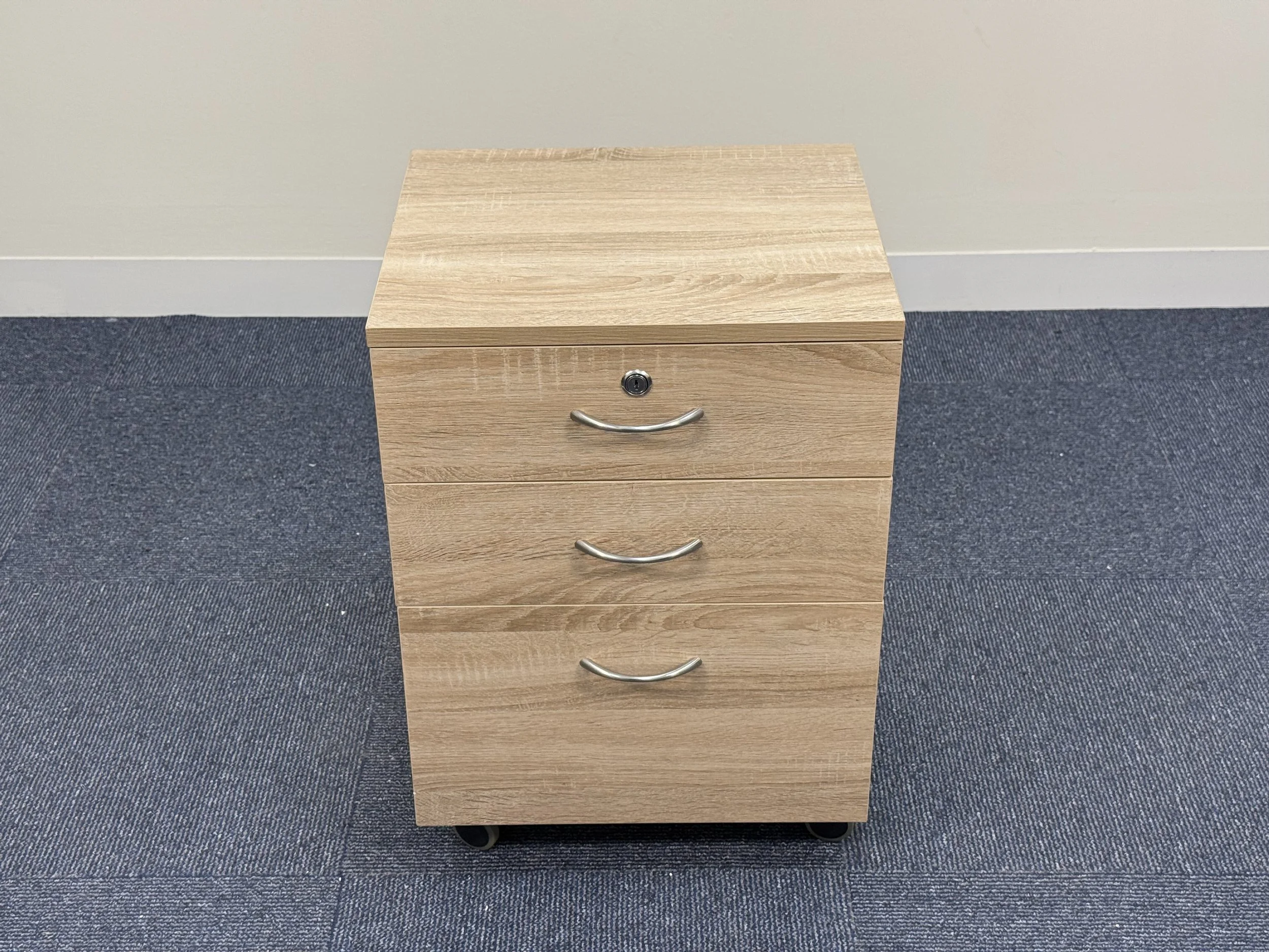 A wooden filing cabinet with three drawers, each with a curved silver handle, placed on a blue carpet in front of a light-colored wall.