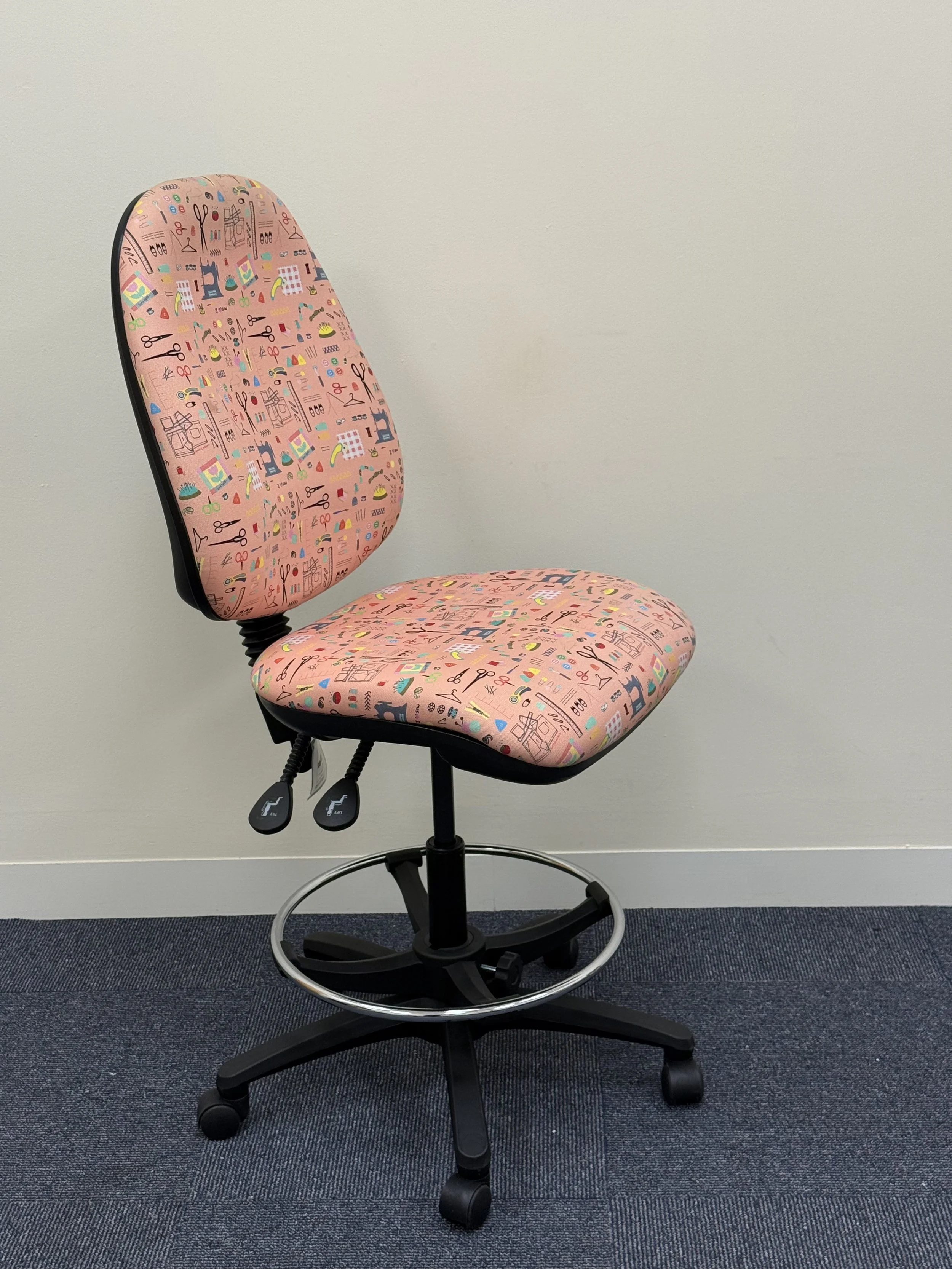 Pink office chair with a colorful pattern of sewing tools and supplies, positioned against a plain white wall, on a grey carpeted floor.