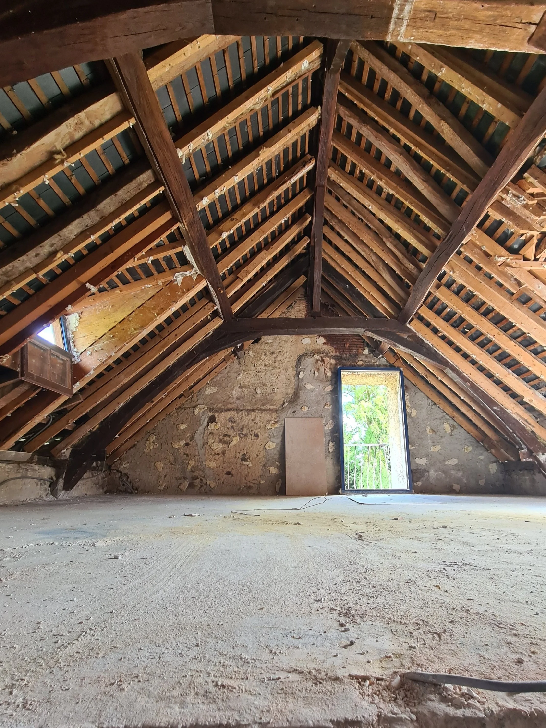 Photo de l'intérieur d'une maison, on voit toutes les poutres, sous les combles, les murs on été retirés, au fond une grande fenêtre laisse entrer la lumière du soleil.