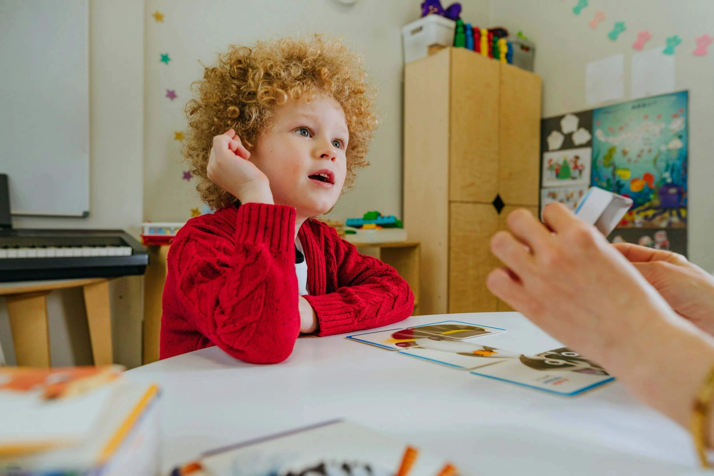 Child with curly hair and red sweater sitting at a table in a classroom, engaging in activity with another person who is holding cards or a book. Background includes a piano, poster, and colorful decorations.