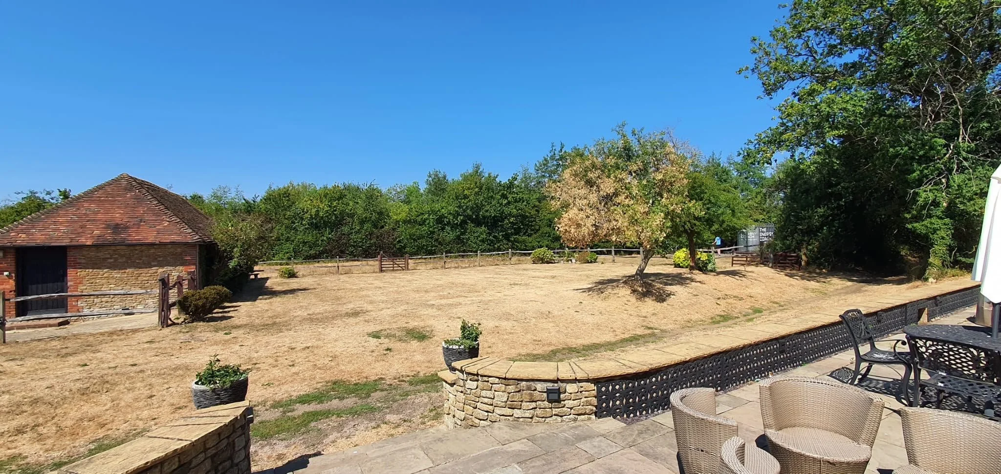 A backyard with a small brick building, a dry grassy yard, several trees, and a patio area with wicker chairs and a black metal table under a white umbrella.