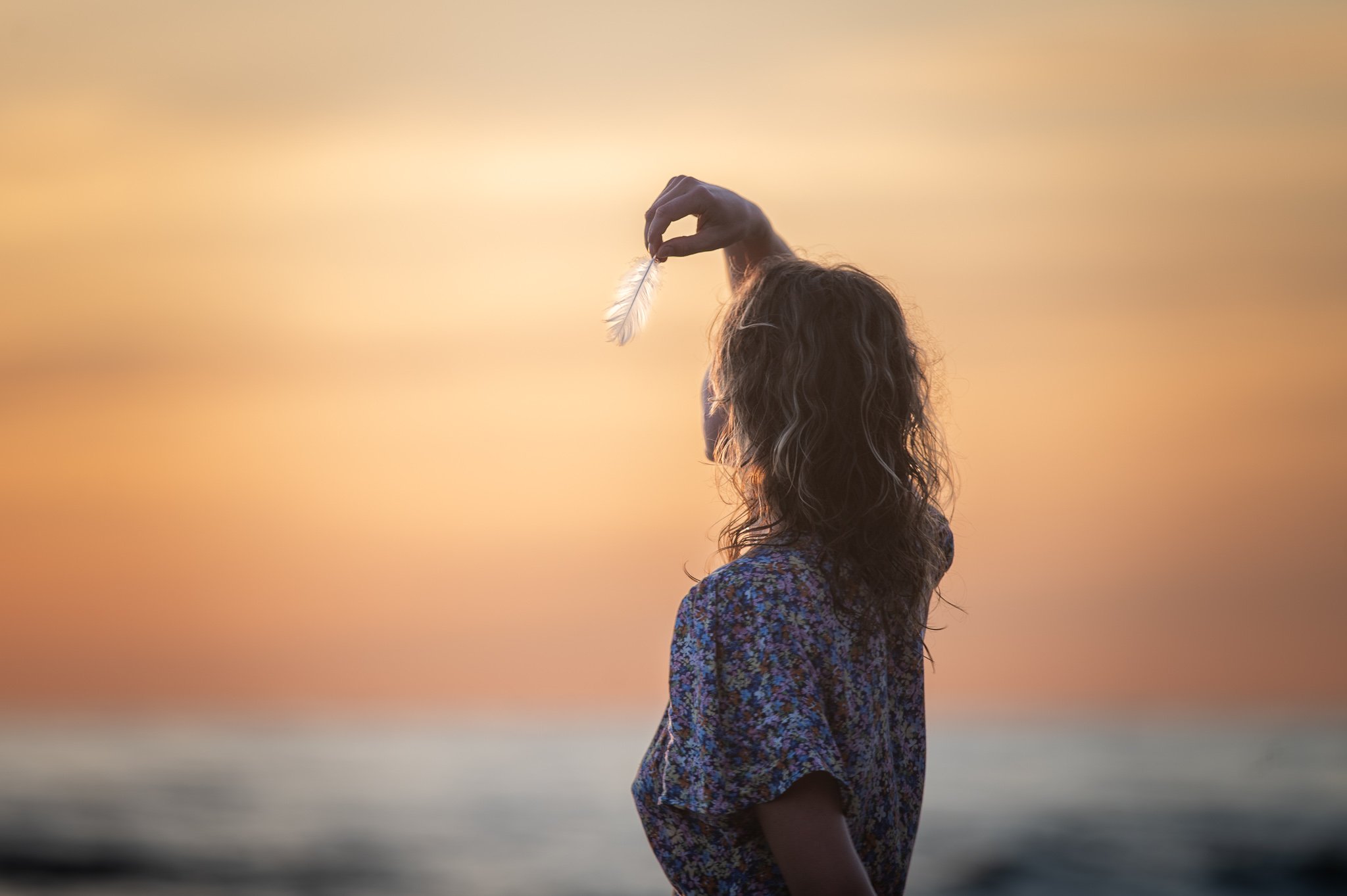 Moters siluetas laikantis plunksną prieš saulėlydį jūros fone – fotografinė metamorfozė moterims, įkvepianti gamtos grožį.