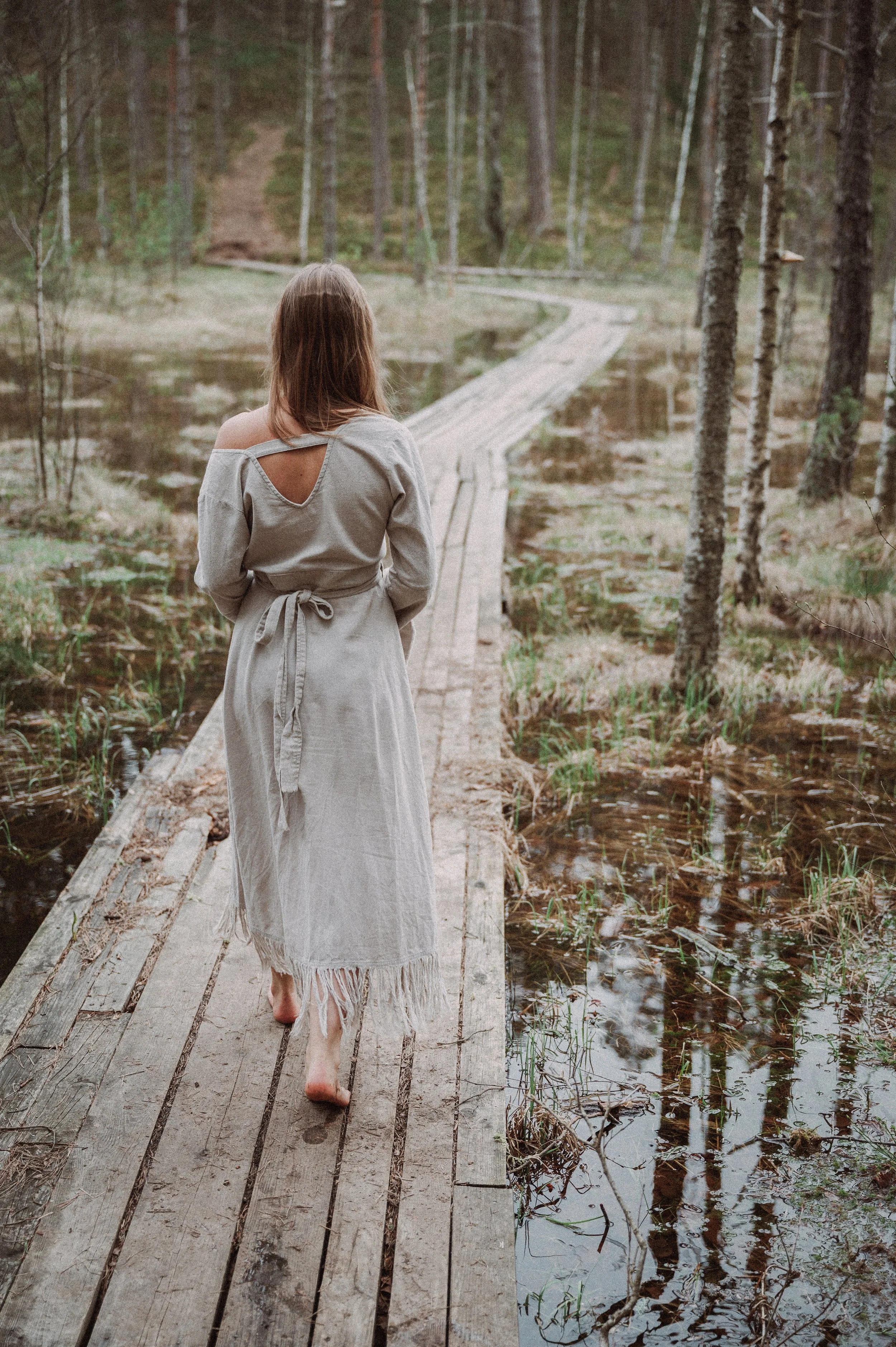 Moteris, pasipuošusi ilga suknele, eina basomis mediniu taku pelkės miško aplinkoje – fotografinė metamorfozė moterims.