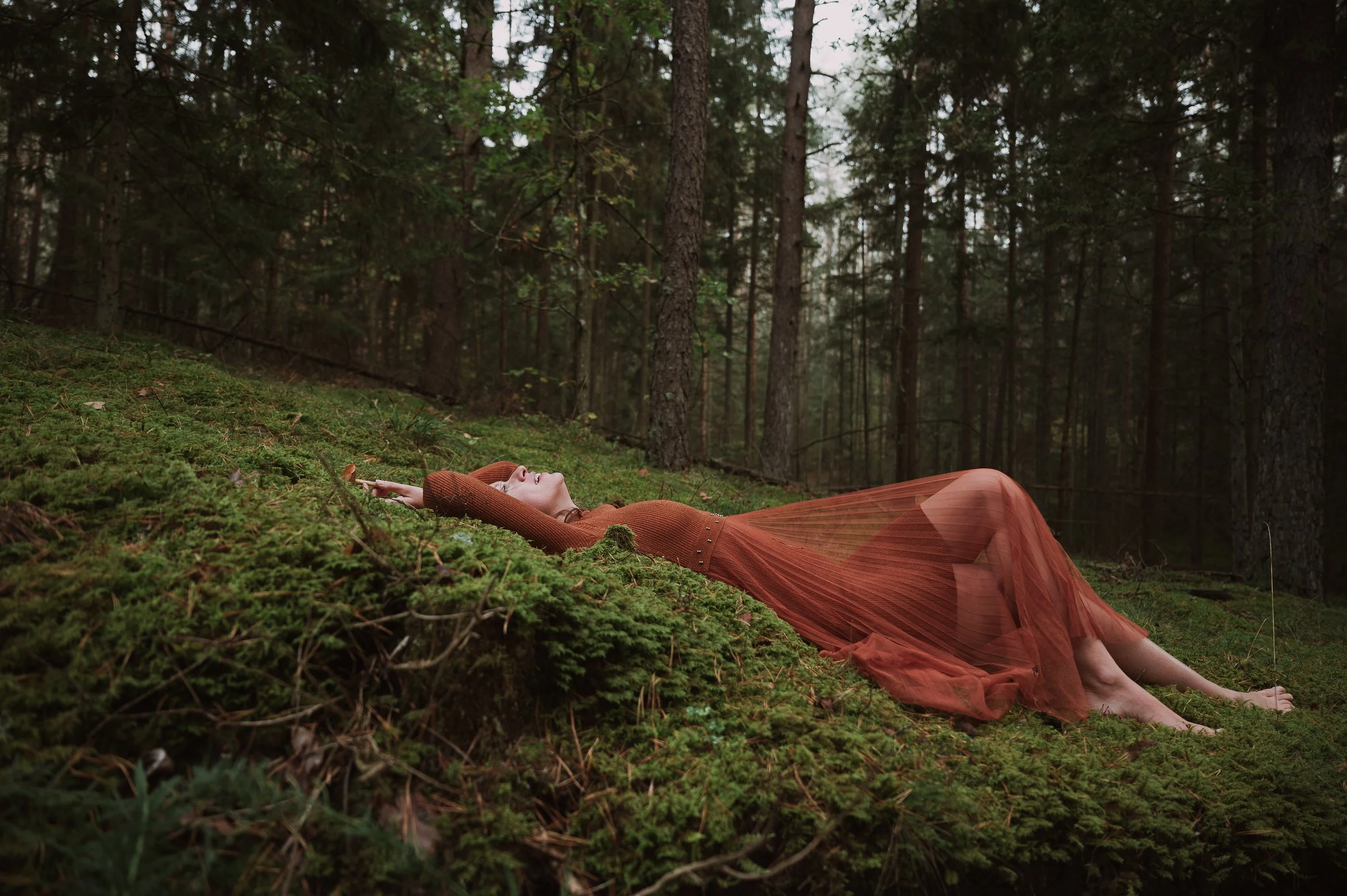 Moteris su permatoma oranžine suknele guli ant samanų miške, apsupta medžių – meninė fotosesija gamtoje.