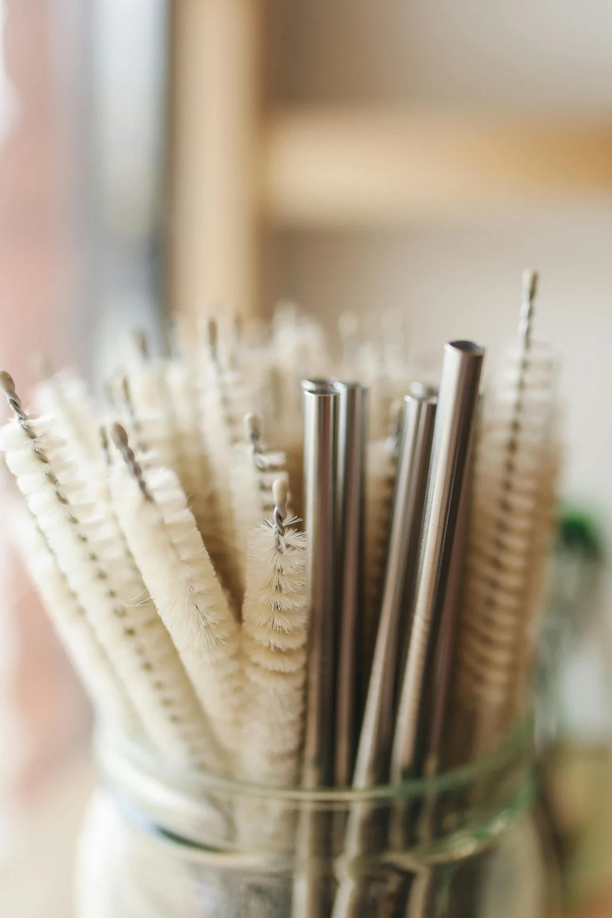 Glass jar packed with reusable stainless-steel straws and cream bottle-brush cleaners, shot in soft natural light.