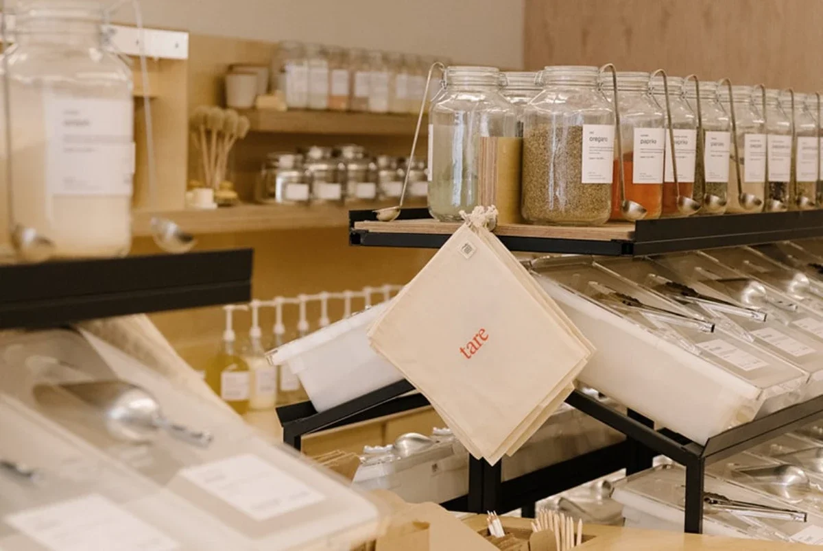 Row of labelled glass spice jars with metal scoops in a refill shop; a cloth “tare” produce bag hangs prominently in front.