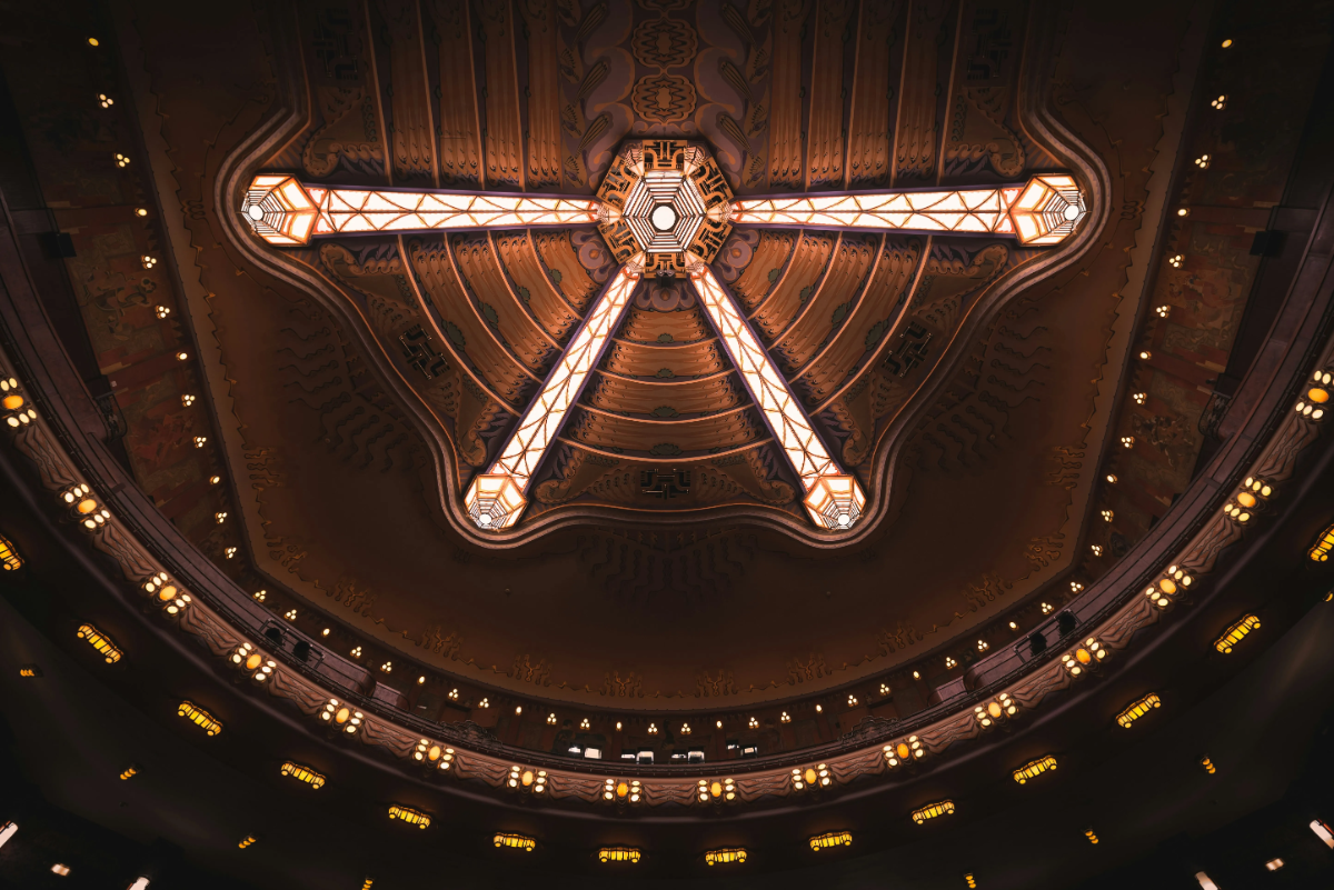 Geometric art-deco chandelier of three illuminated beams radiates from ornate theater ceiling, surrounded by balcony lights (concept image, not Orient Express Corinthian)..