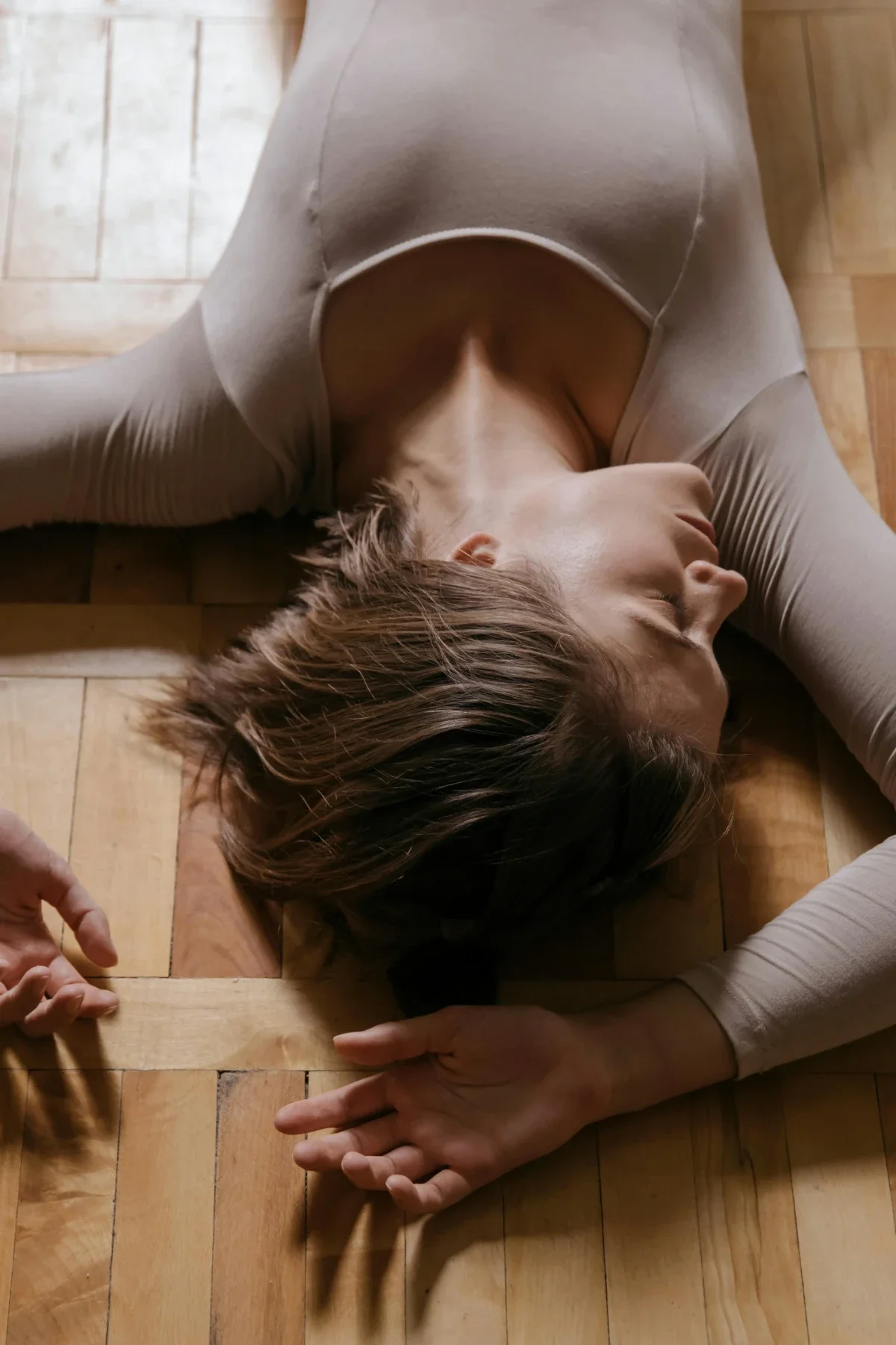  Dancer in beige leotard lies sprawled on a polished parquet floor, eyes closed and hands resting softly by her sides. 