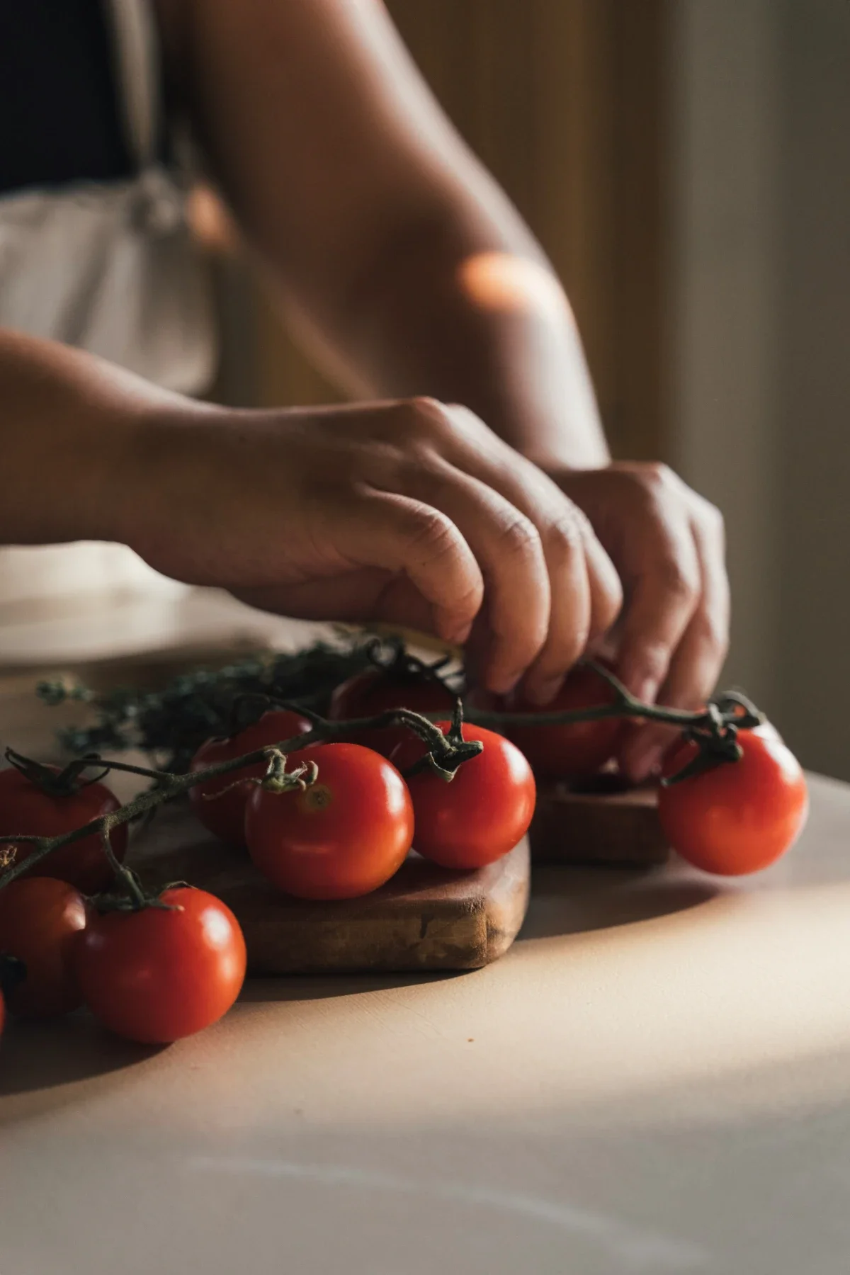 Cook’s fingers arrange ripe cherry tomatoes on the vine atop a wooden board in soft afternoon light.