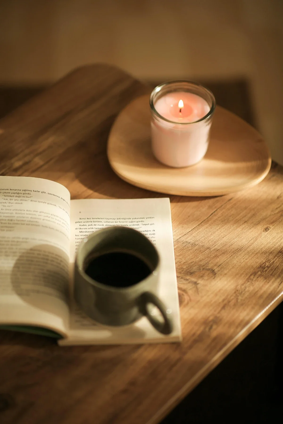 Pink jar candle next to an open book and a mug of black coffee on a warm wooden table, creating a cozy reading nook.