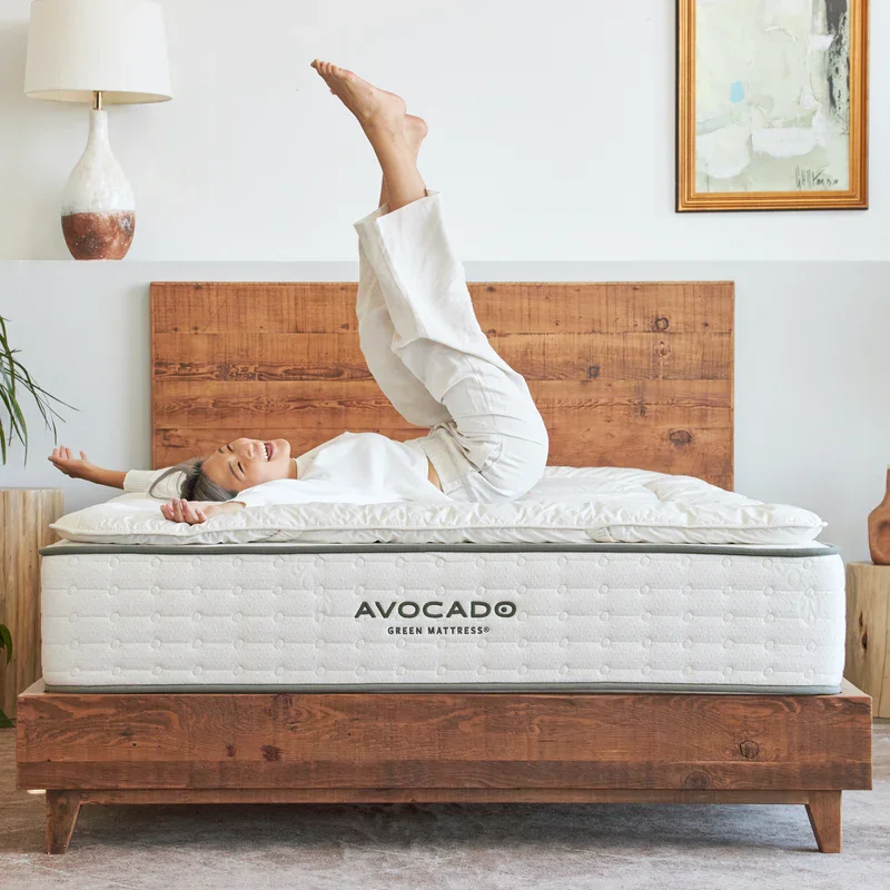 Smiling woman on Avocado plush hotel topper atop Green Mattress, legs playfully raised toward ceiling.
