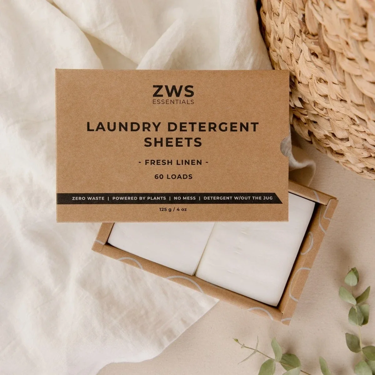  Kraft box of “ZWS Essentials” fresh-linen laundry detergent sheets opened on soft fabric beside a woven hamper. 
