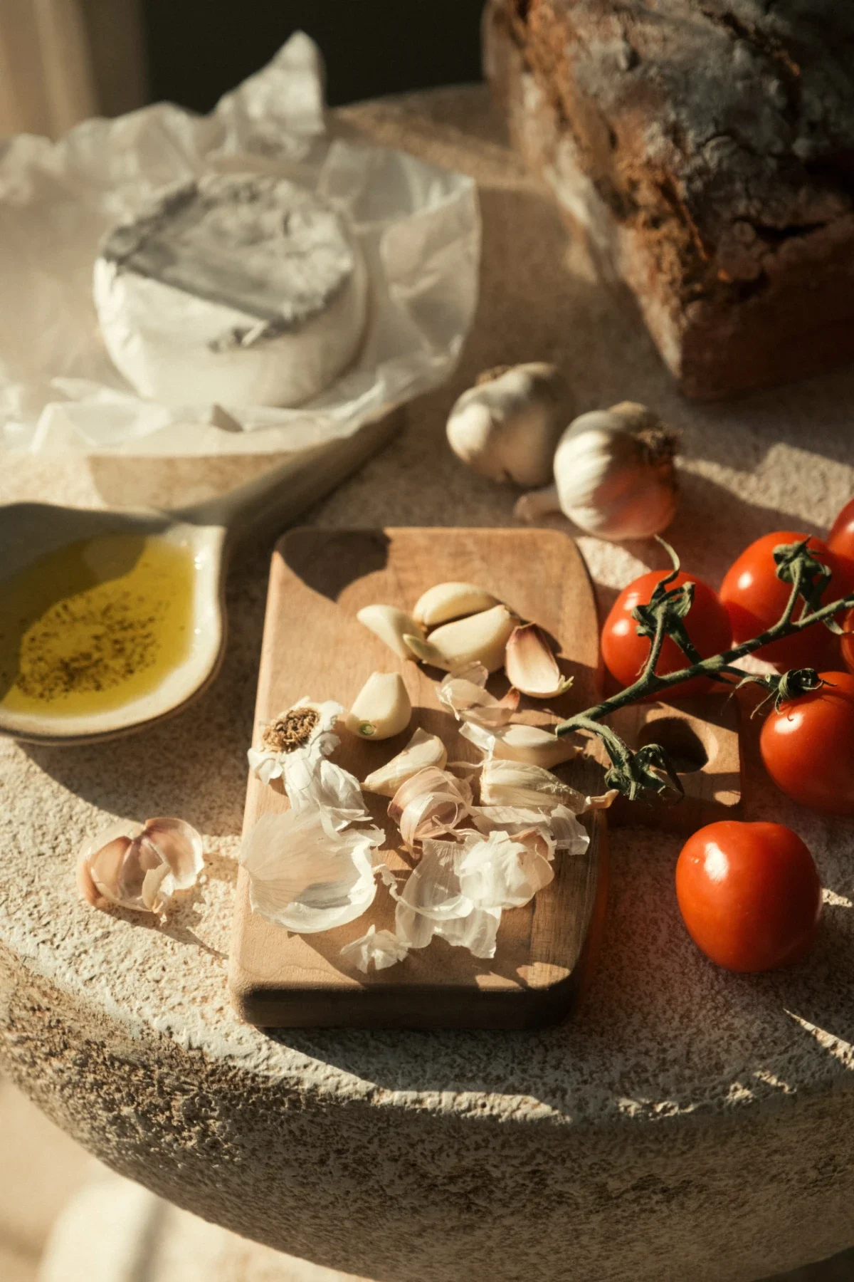 Garlic cloves, vine tomatoes, brie, olive-oil dish and crusty bread on a sunlit stone table, warm, old-world kitchen vibe.