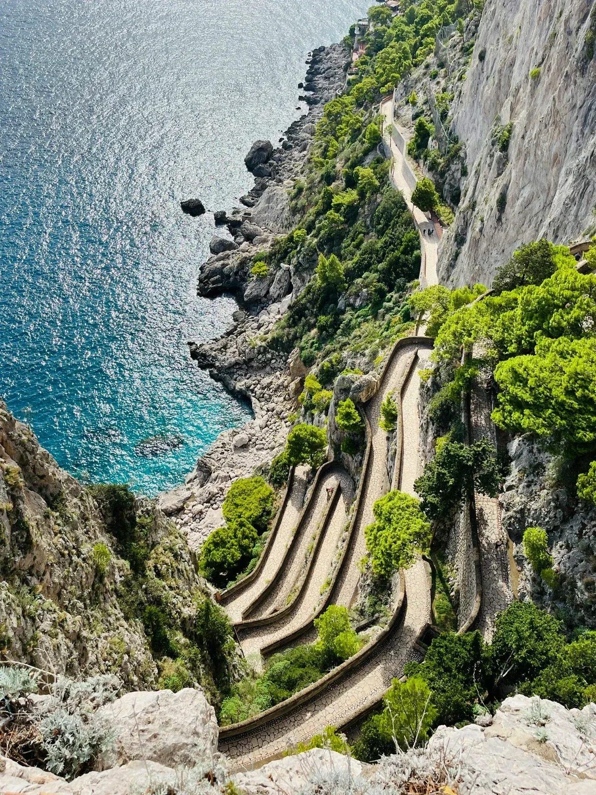  Aerial view of serpentine Via Krupp descending Capri’s limestone cliffs beside sparkling turquoise water on Capri. 