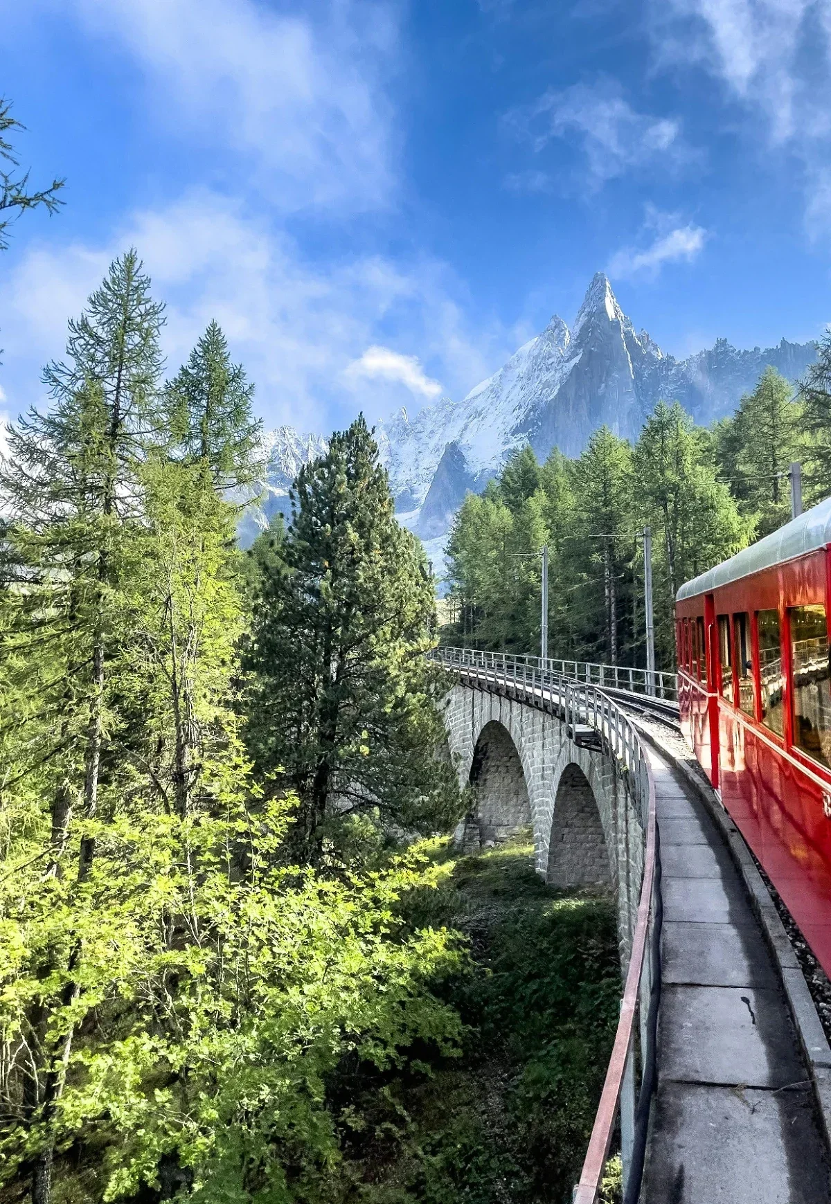 Red alpine train crossing stone bridge through forest with towering snow-covered mountains in the background.