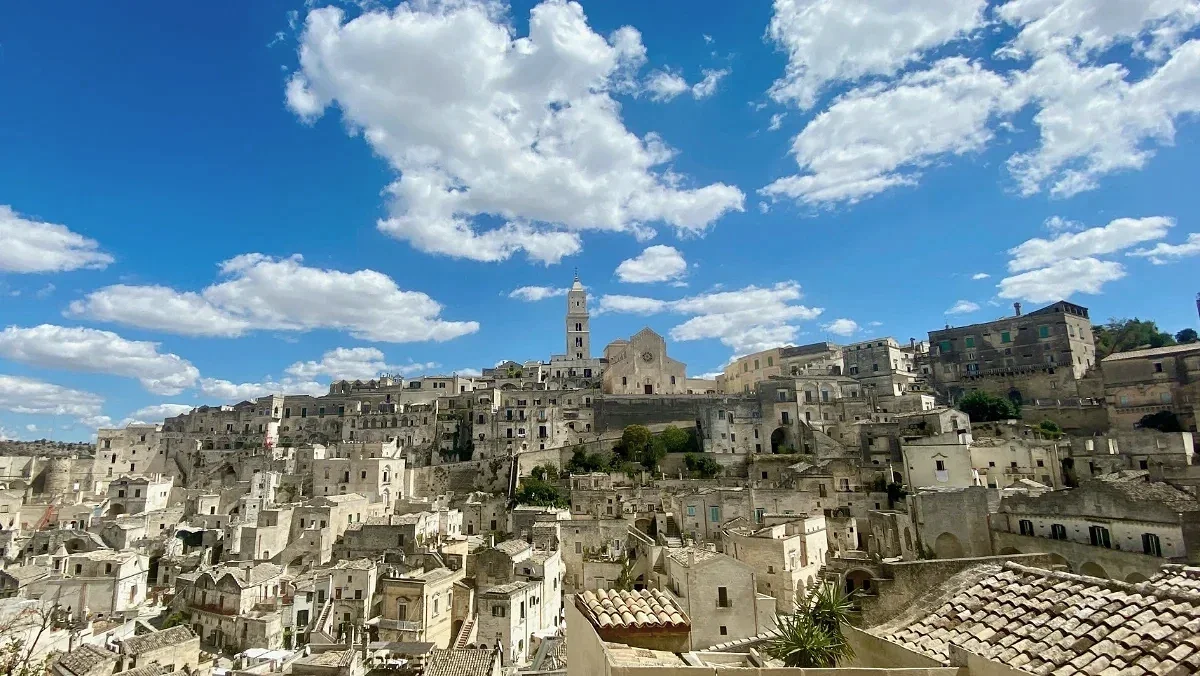  Sunlit stone Sassi cave homes and cathedral tower shape Matera skyline beneath a vivid blue sky. 