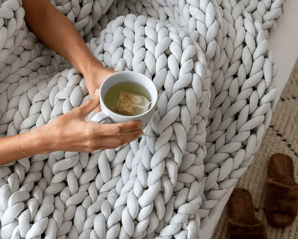 Woman relaxing with herbal tea wrapped in soft-grey Gravity chunky-knit weighted blanket on sofa.
