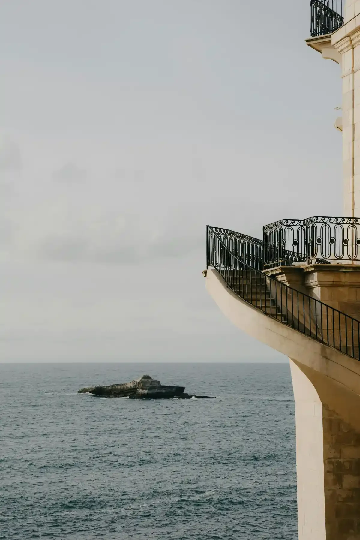 Curved limestone balcony with ornate black railing overlooking the sea and a solitary offshore rock.