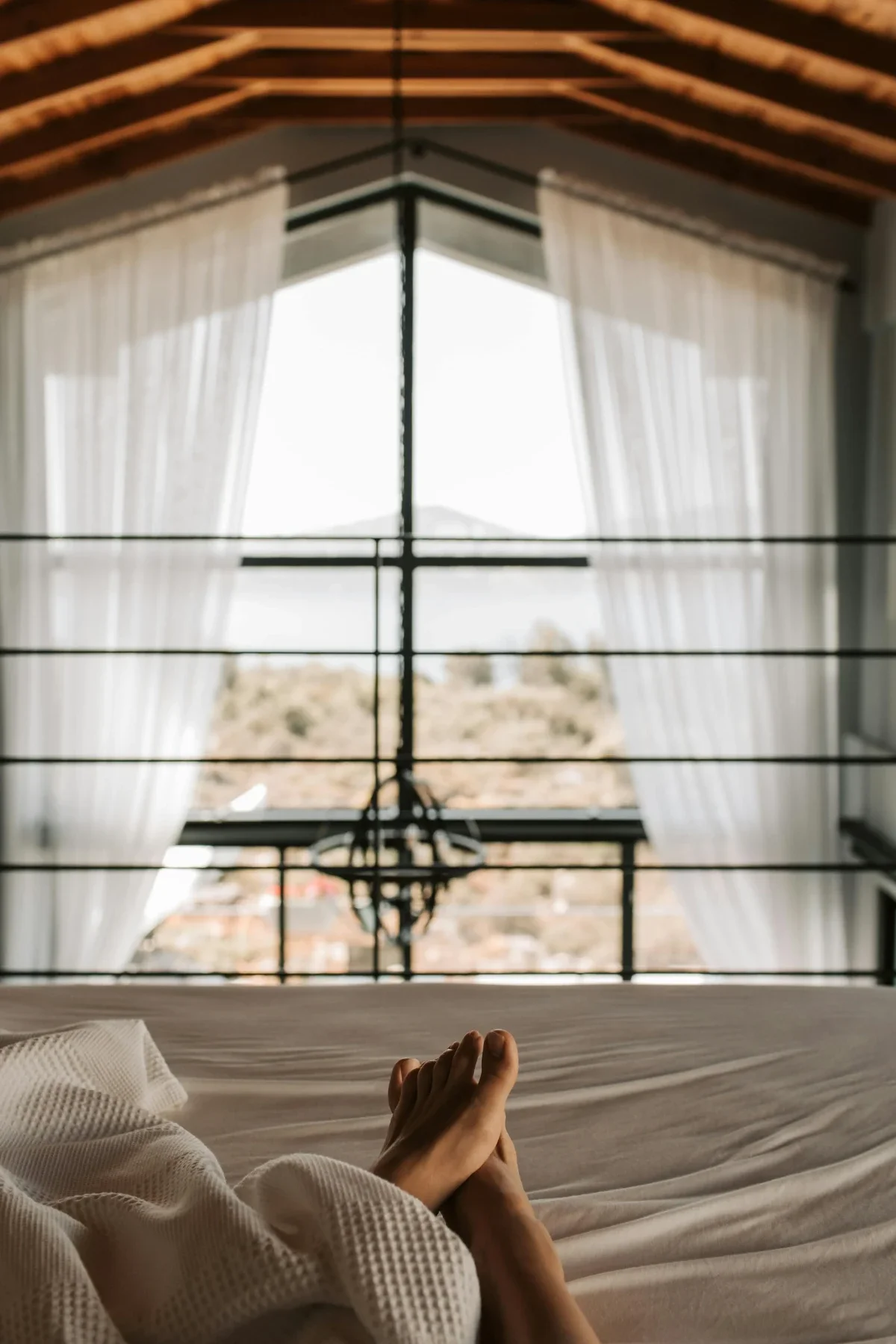 Relaxed view from bed in a lofted cabin, bare feet crossed toward tall window with sheer curtains and distant hills.