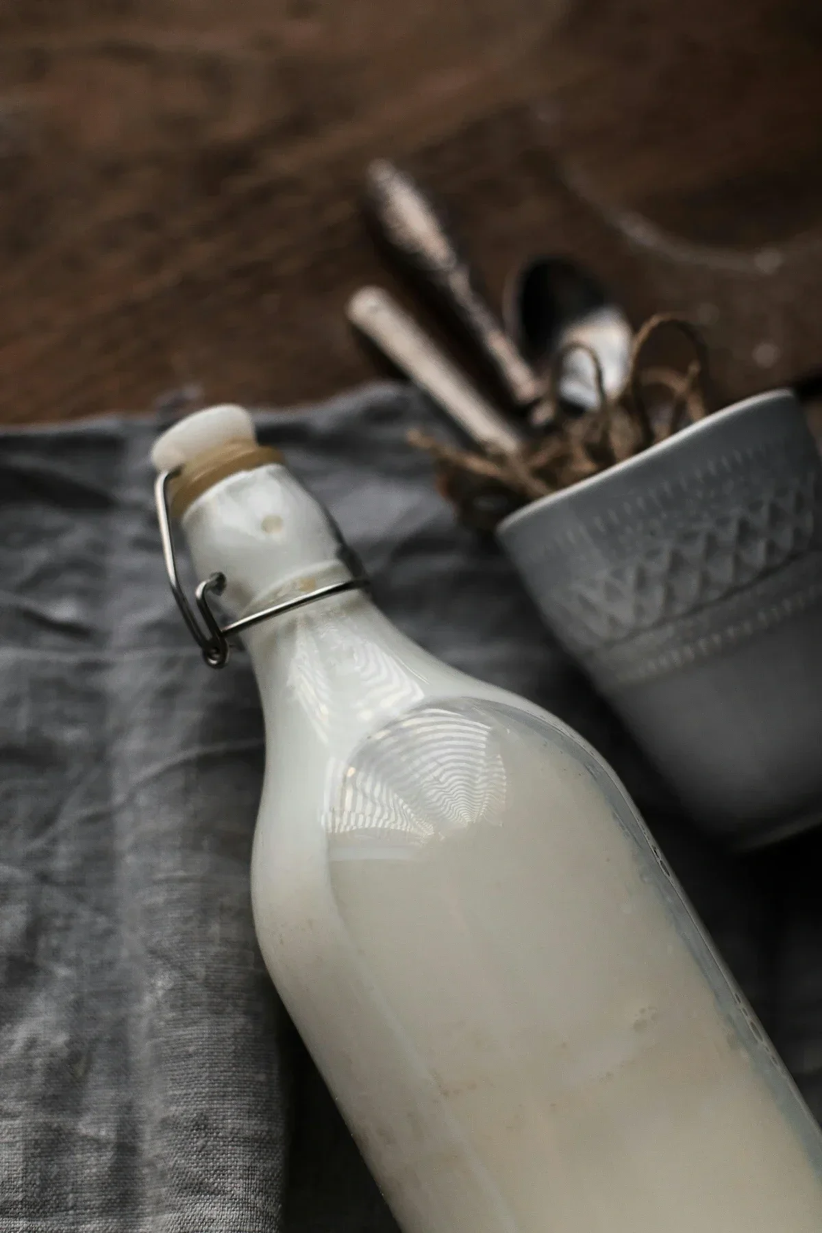 Bottle of homemade plant-based milk on cloth with utensils, soft natural light and minimal kitchen styling.