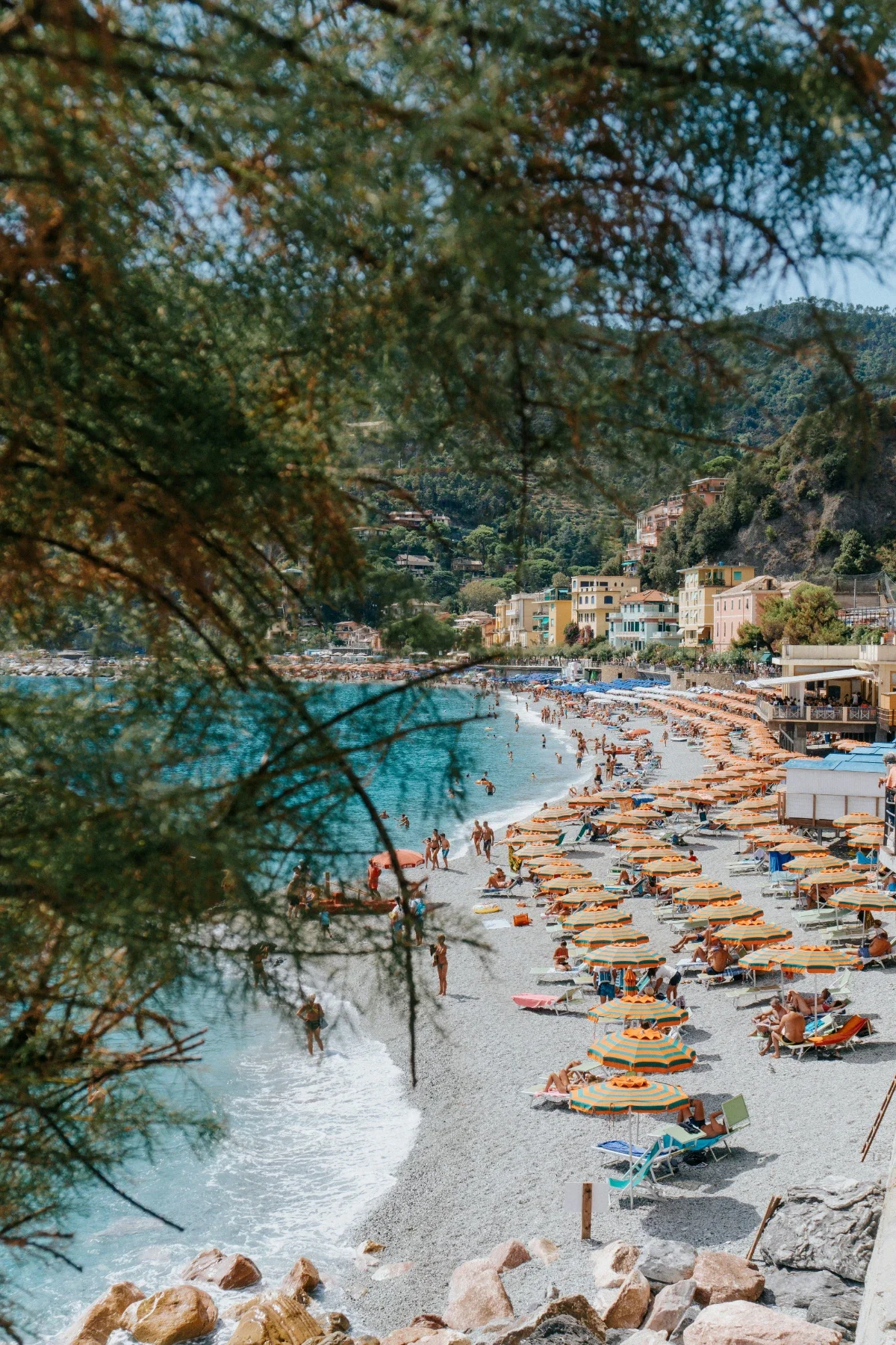  Rows of orange-yellow umbrellas line busy pebble shore at Monterosso al Mare with green hills beyond. 