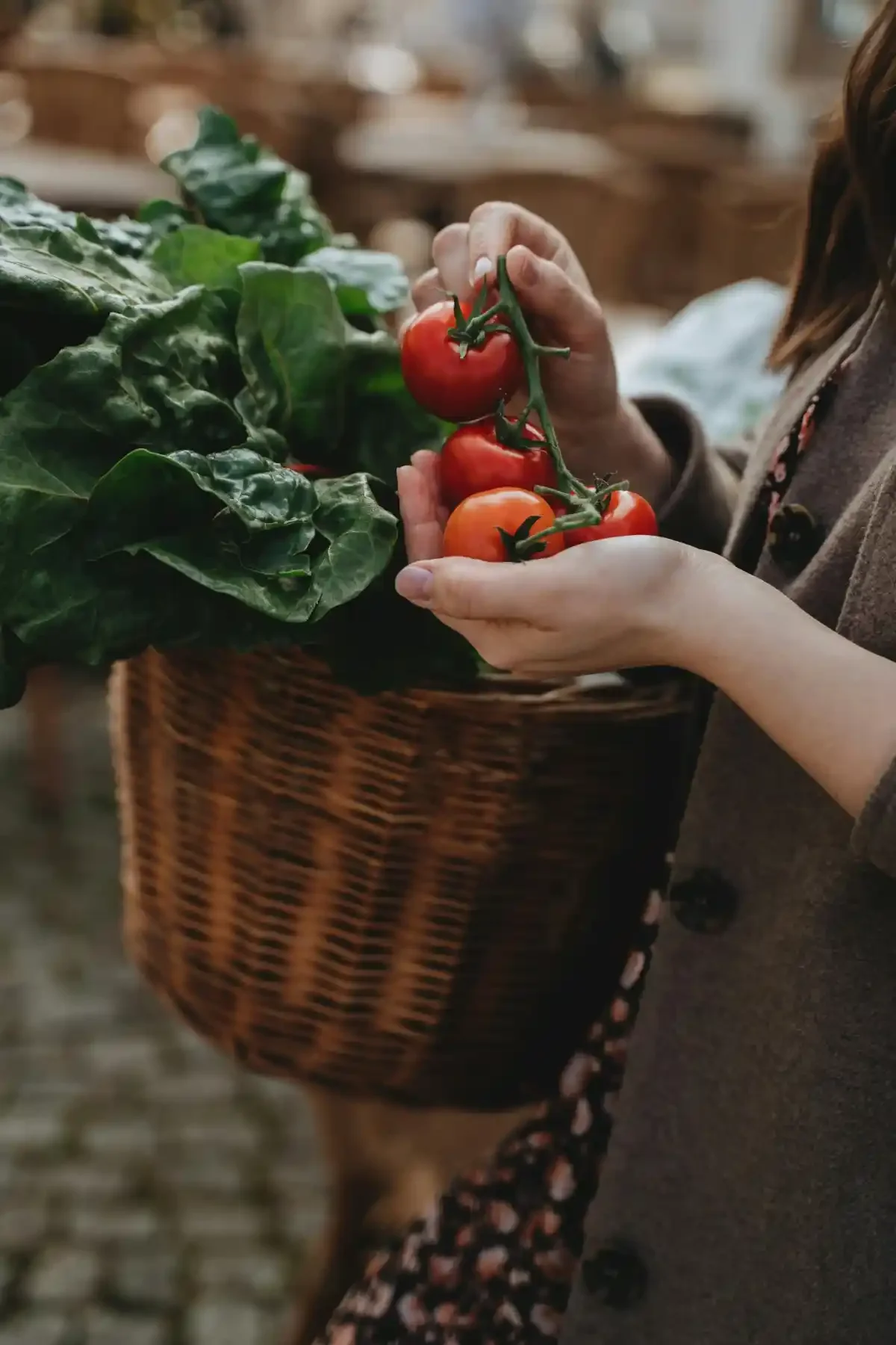 Shopper holding vine-ripened tomatoes above a basket brimming with leafy greens.