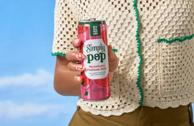 Person in crochet top holds a tall pink Simply Pop Strawberry Prebiotic Soda can against a bright blue sky.