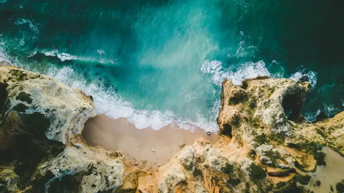 Aerial view of Praia da Balança: aquamarine Atlantic swells curl onto a narrow sand strip framed by limestone cliffs near Lagos.