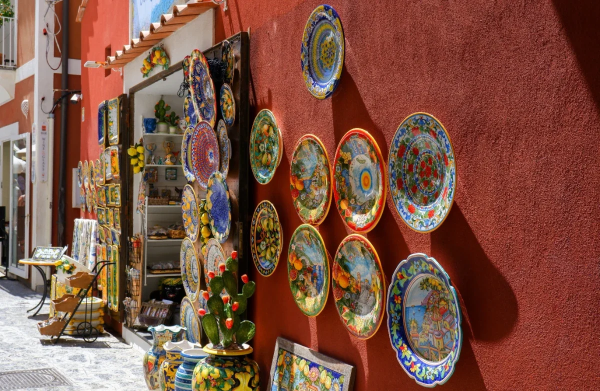 Sunlit ceramics shop with vivid hand-painted plates and lemons on a red stucco wall in the Amalfi Coast.