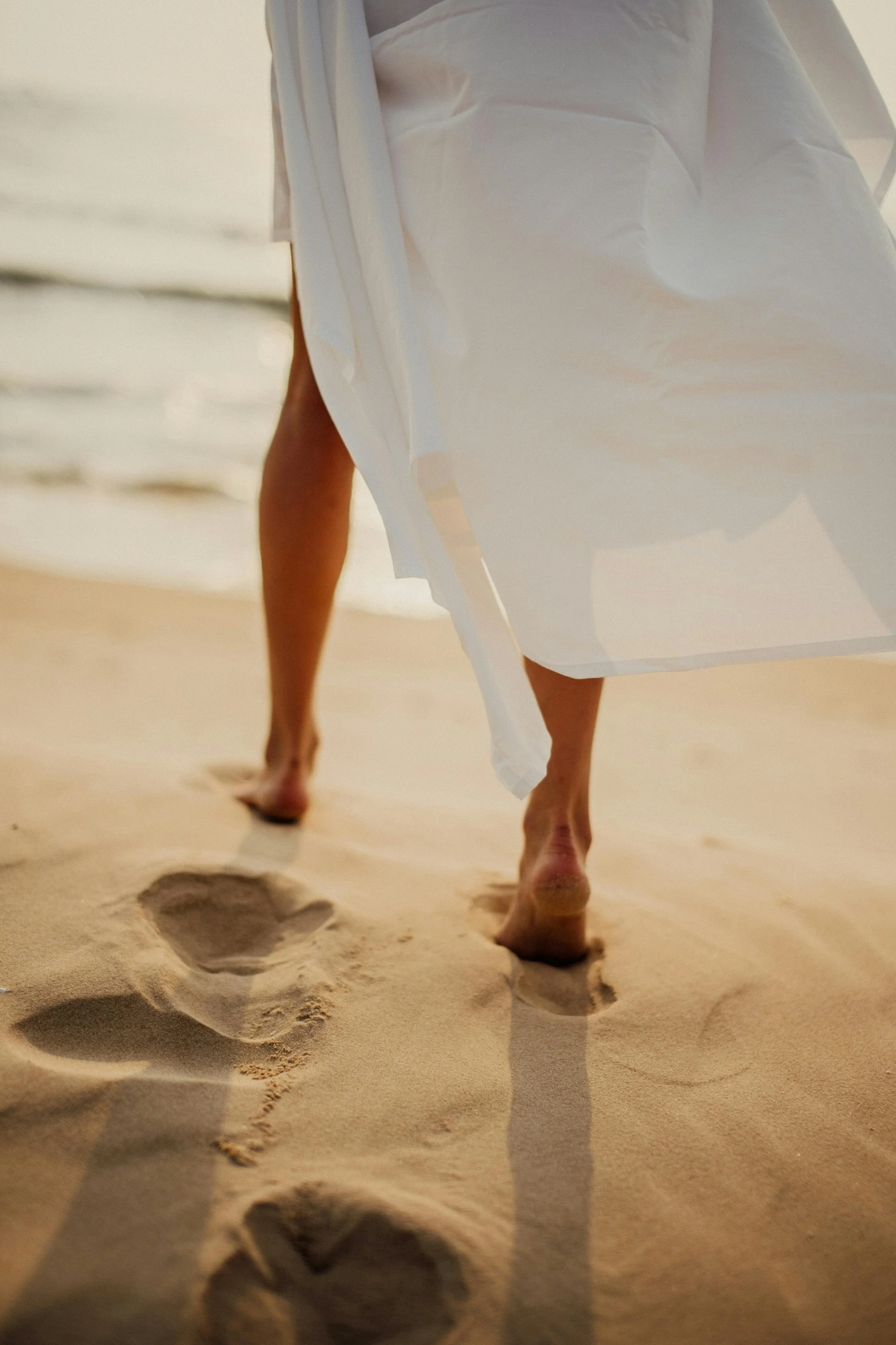 Close-up of a woman in a flowing white dress walking barefoot on a sandy beach, leaving fresh footprints near the shoreline.