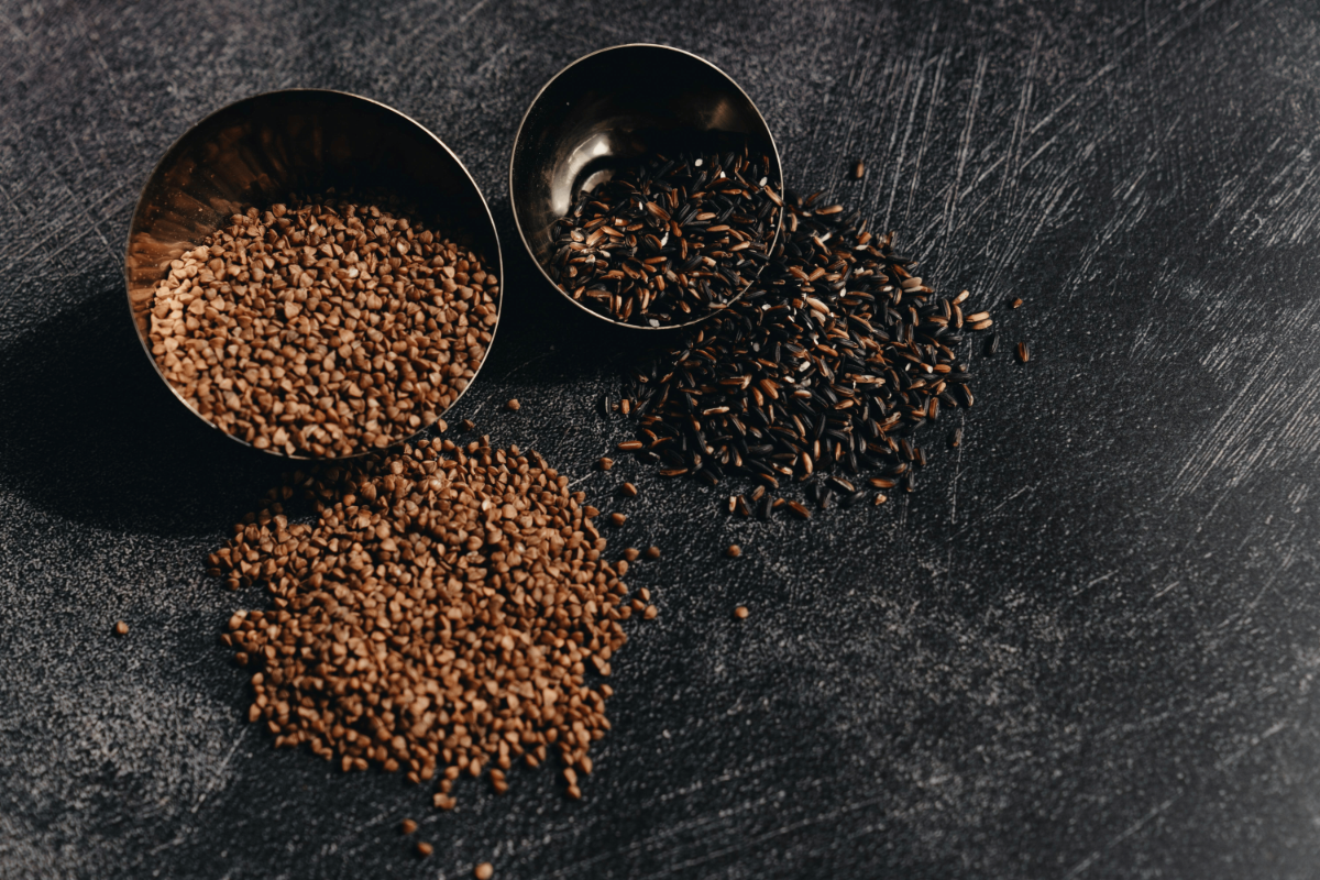 Metal bowls of raw buckwheat and wild rice spilling onto textured black surface, illustrating contrasting whole-grain types.