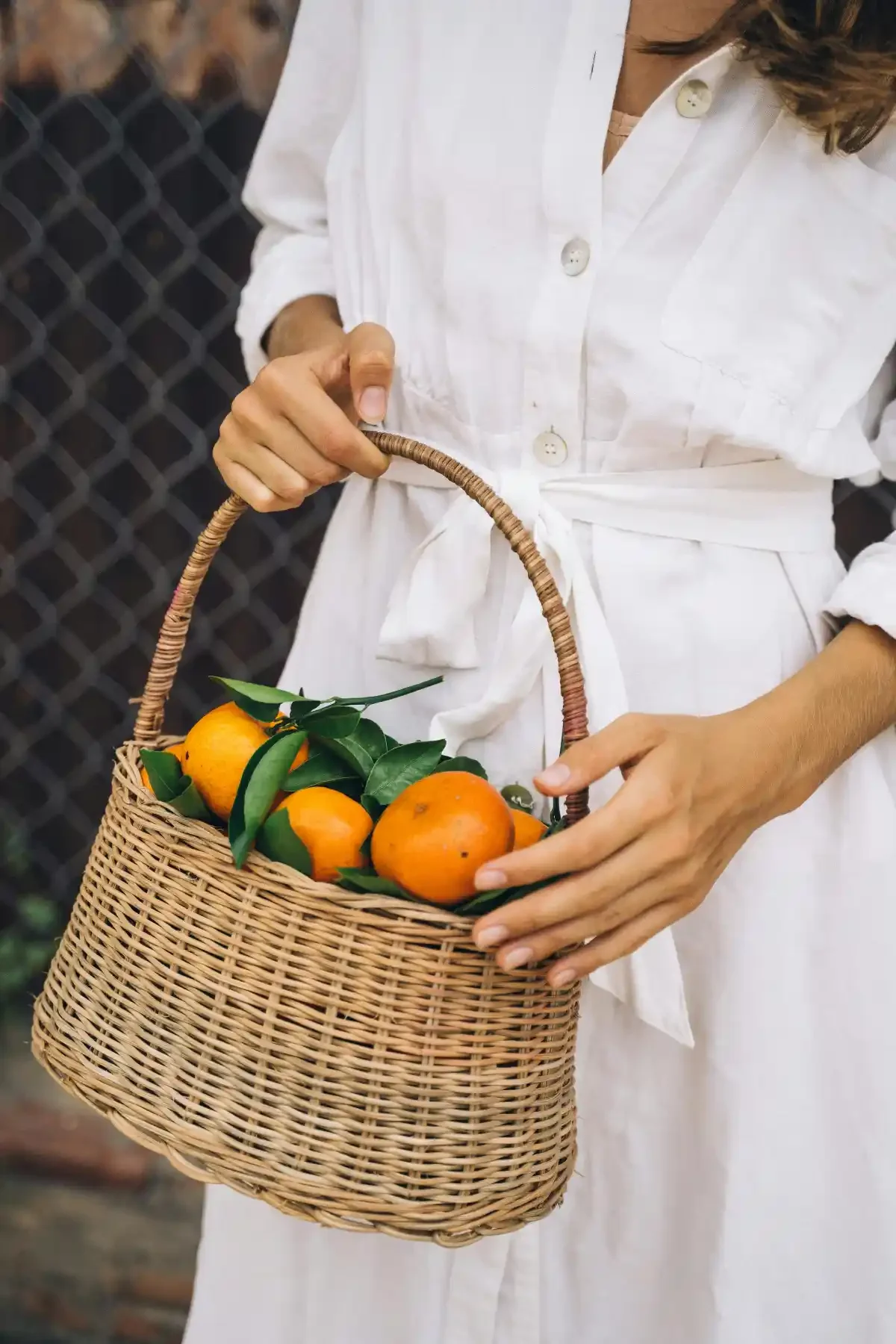 Wicker basket of bright oranges with leaves, carried by a woman in a white dress.