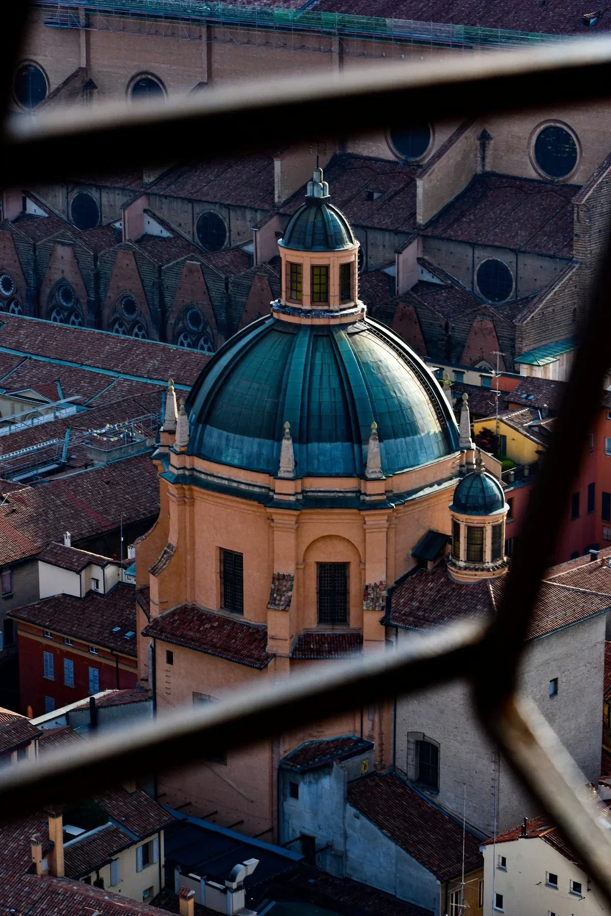  Latticed viewpoint over terracotta rooftops to a teal-green church dome crowning historic Bologna. 