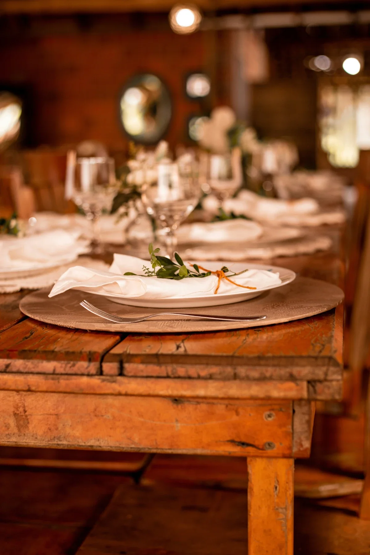 White plate with folded linen napkin and green sprig set on a weathered wooden dinner table.
