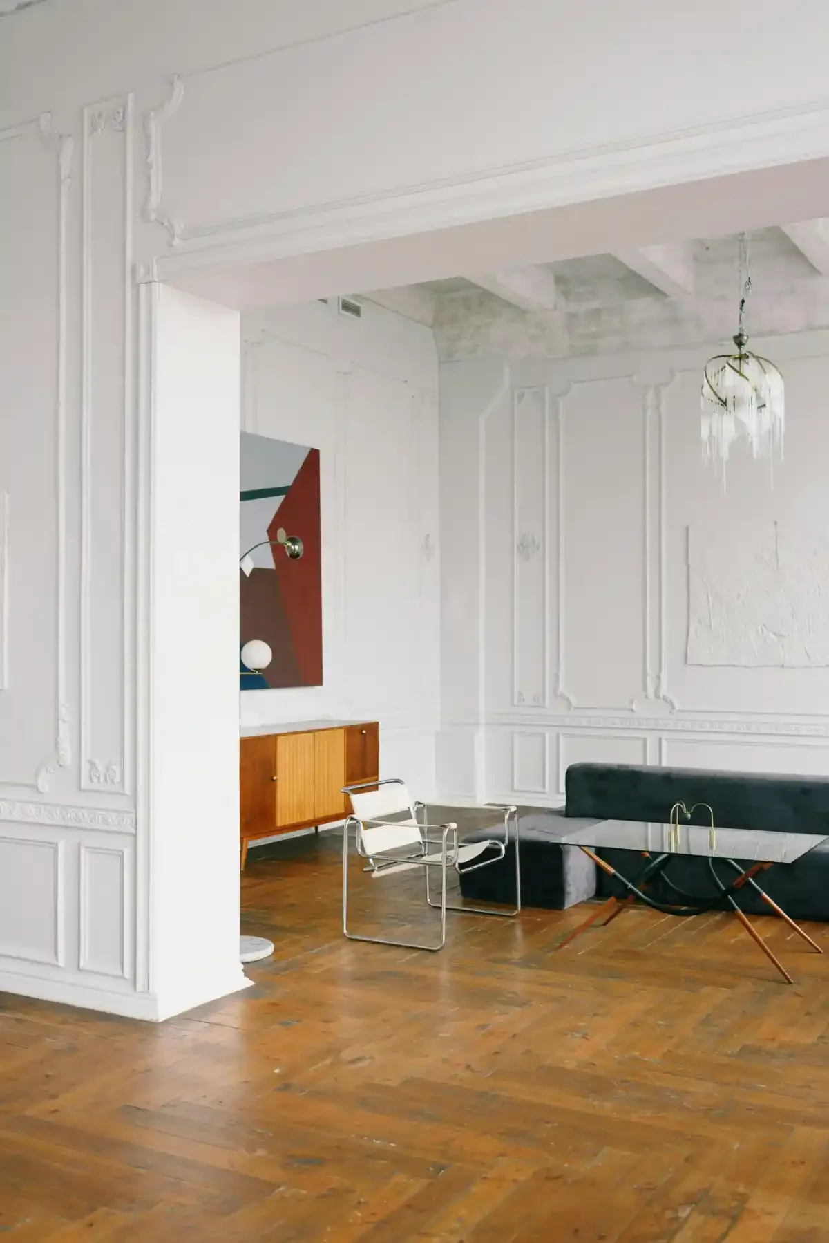 Bright eclectic living room featuring white Wassily chair, mid-century cabinet, moulded walls and parquet flooring.