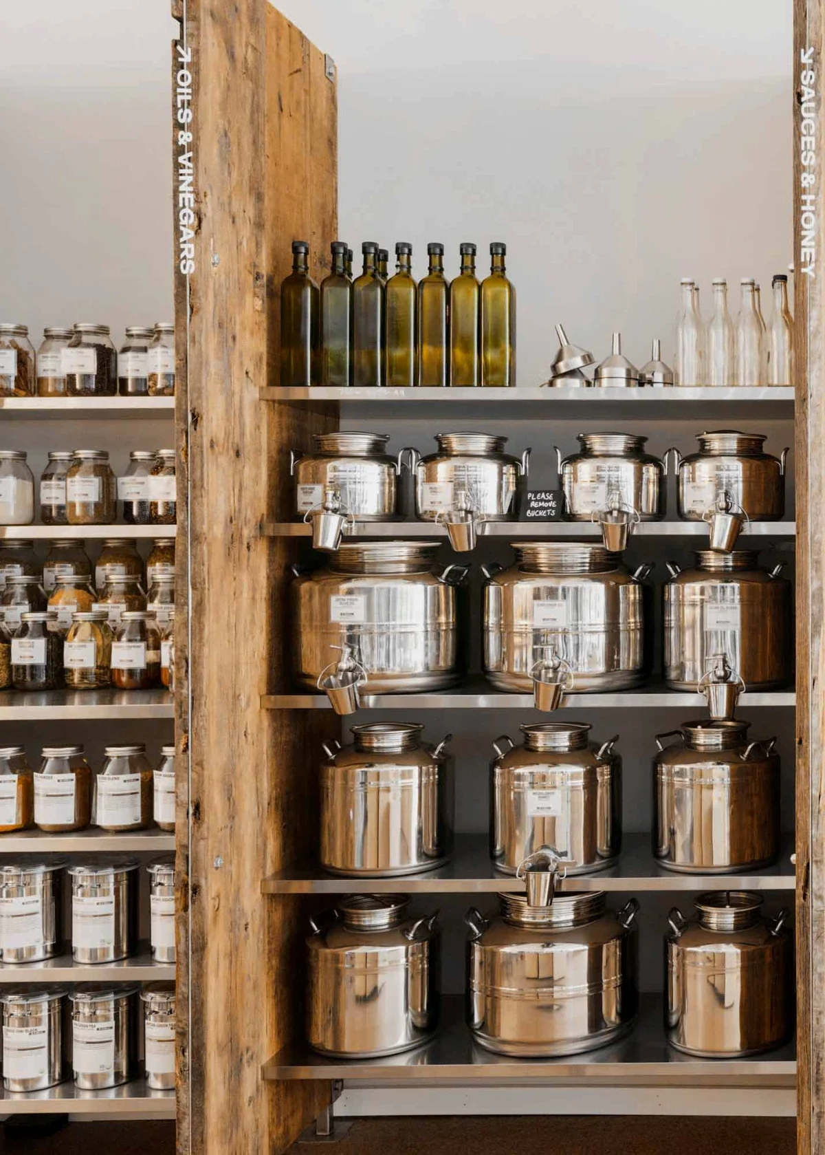 Rows of stainless fusti tanks and refillable olive-oil bottles on rustic shelves in a zero-waste store.